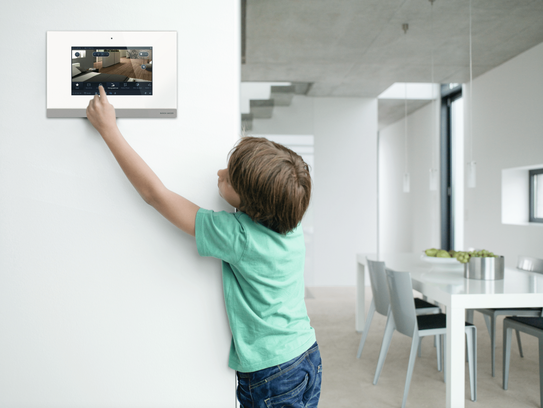ABB - Busch-Jäger zeigt einen kleinen Jungen, welcher an Wandsteuerung mit Kamera tippt im Esszimmer.
