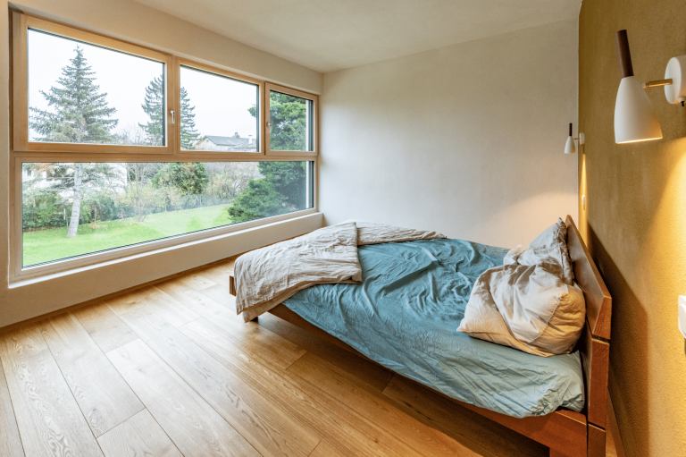 AL Architekt ZT zeigt ein kleines Schlafzimmer mit einem Doppelbett aus Holz, weissen Wandleuchten und einem großem Panorama-Fenster mit Ausblick ins Grüne.