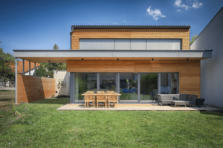 AL Architekt ZT zeigt ein helles Einfamilienhaus aus Holz mit Flachdach und einer überdachten Terrasse mit Sitzgruppe aus Rattan, einem Carport und grauen Rollos.