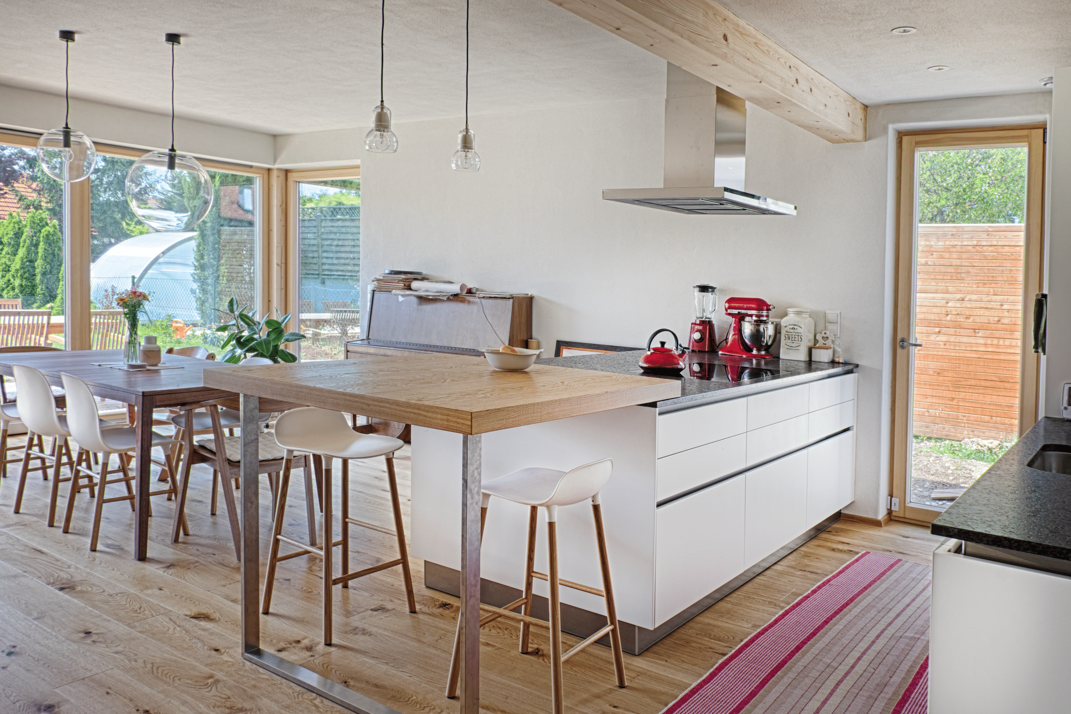 AL Architekt ZT zeigt eine Küche aus hellem Holz und weissem Küchenblock mit einer Bar und weissen Hockern mit Beinen aus Holz und eine Essgruppe.
