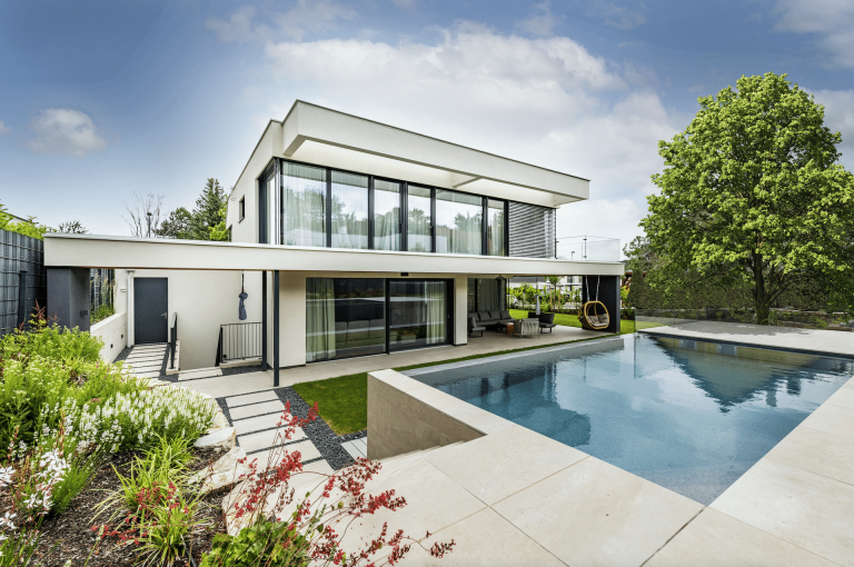 Modernes, helles Flachdach-Wohnhaus mit Glasgeländer-Balkon, großen Glasfronten, überdachter Terrasse und erhöhtem Pool von AL Architekt.