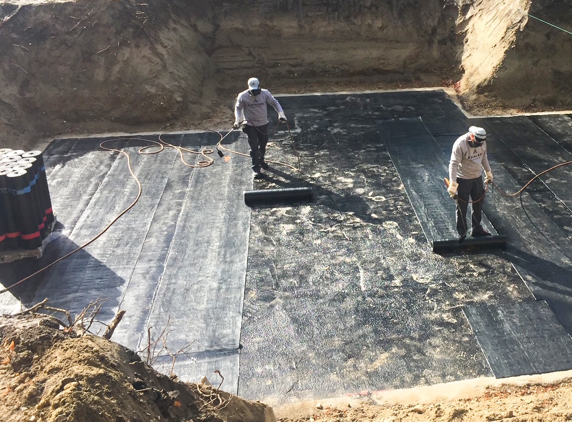 Askalani Abdichtungstechnik zeigt Arbeiter im Aushub einer Baustelle beim Abdichten der bereits erstellten Kellerbodenplatte.