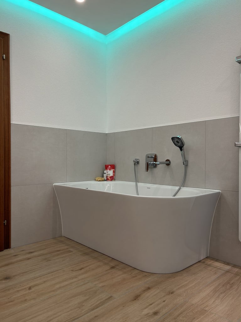 Badezimmer mit großformatigen hellgrauen Fliesen, Laminatboden, Badewanne mit wandmontierter Armatur und herabgesetzter Decke mit Spots und LED-Licht.