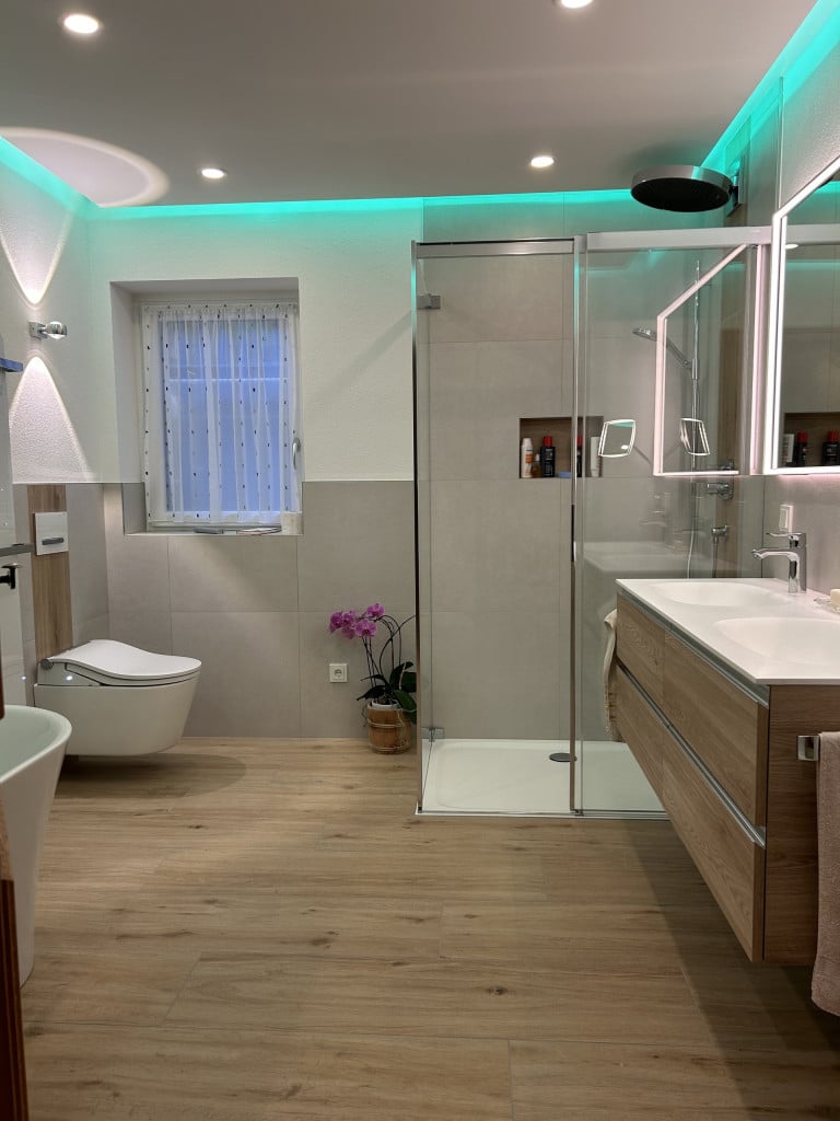 Badezimmer mit großformatigen hellgrauen Fliesen, Duschkabine mit Glasschiebetür und Duschtasse, wandhängender Toilette und herabgesetzter Decke mit Spots und LED-Licht.