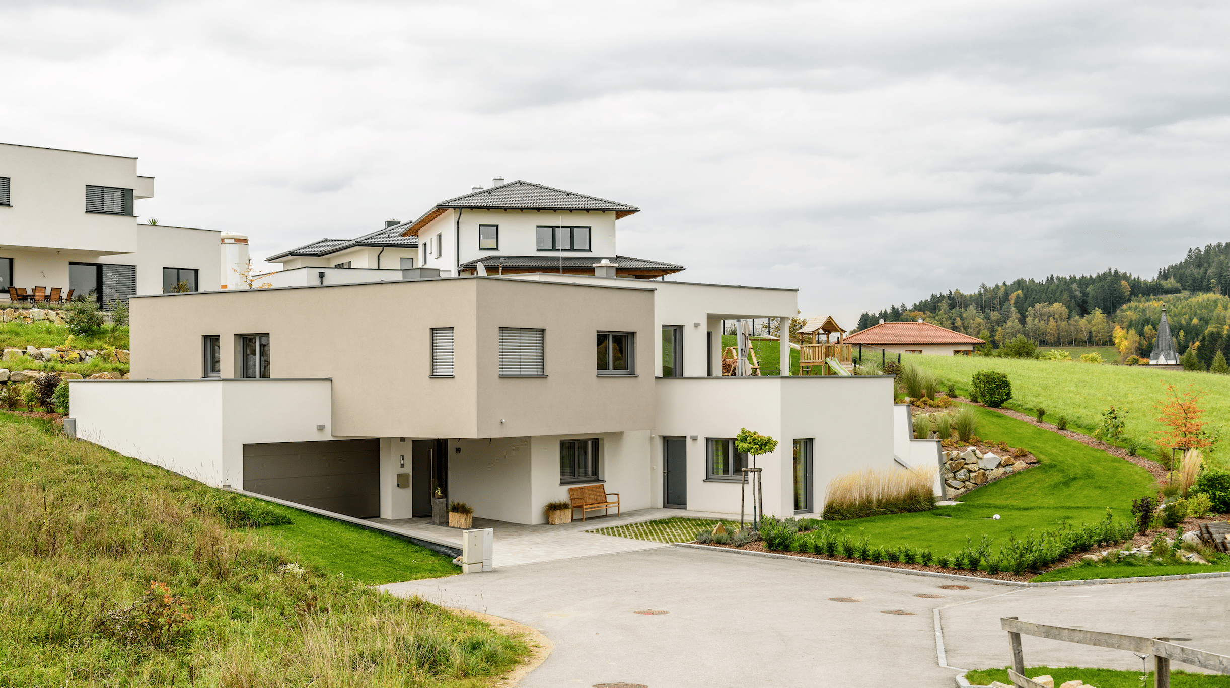 Bad Zeller zeigt ein großes, beige-weißes Einfamilienhaus, mit großer Einfaht, Garage und Flachdach in Hanglage.
