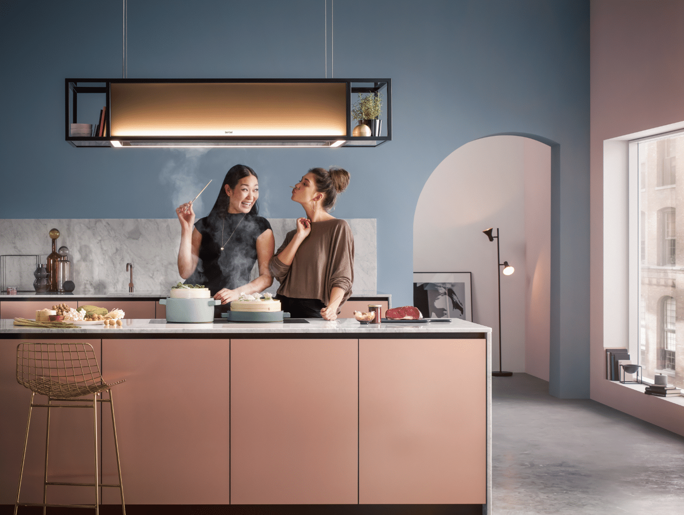 Zwei Frauen sind vergnügt beim Kochen in einer modernen Küche unter Verwendung eines beleuchteten berbel Dunstabzugs mit der Bezeichnung Deckenlifthaube Skyline Frame.