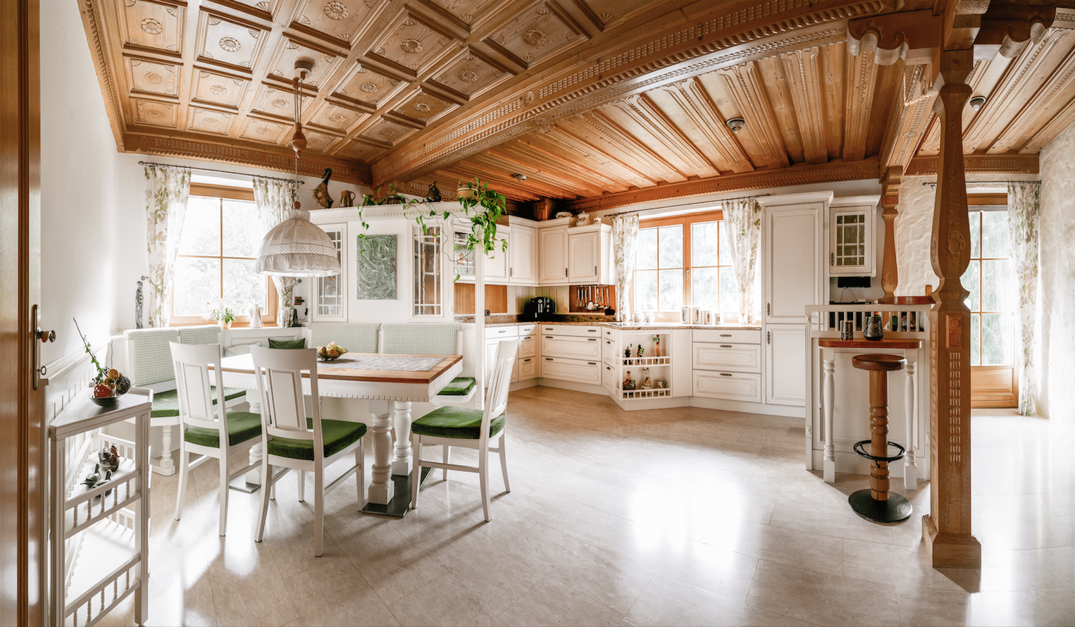 Lichtdurchflutete Küche vom Traditionsunternehmen Böhm Möbel, gefertigt im Landhausstil, mit weißen Einbauschränken, kunstvoll geschnitzten Holzsäulen, traditioneller Holzdecke und einem Esstisch mit Eckbank und Sesseln.