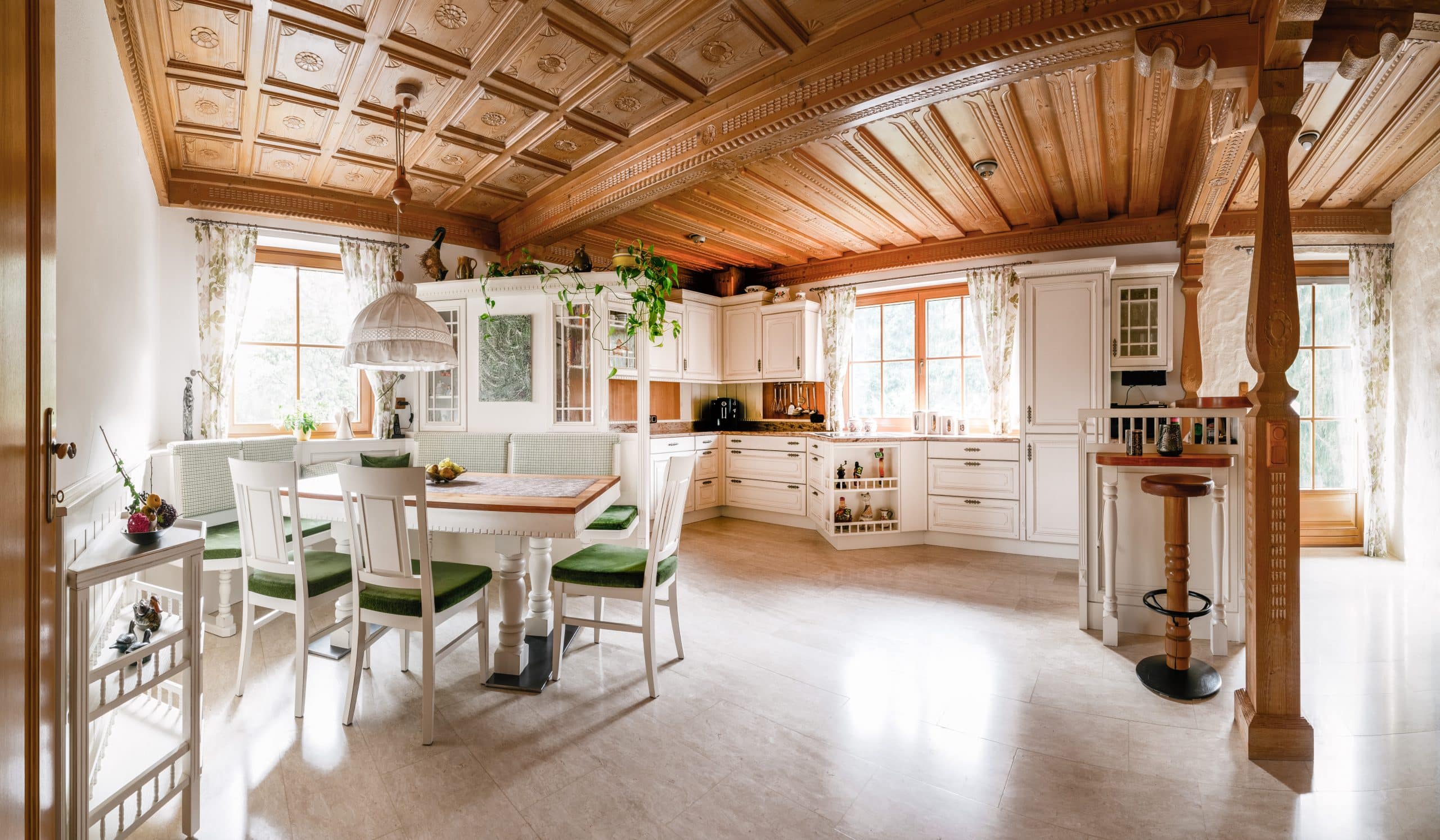 BÖHM MÖBEL zeigt eine weiße Küche im Landhausstil mit verspielten Schnitzereien, brauner Arbeitsplatte und einen Esstisch mit Stühlen und grünen Sitzkissen.