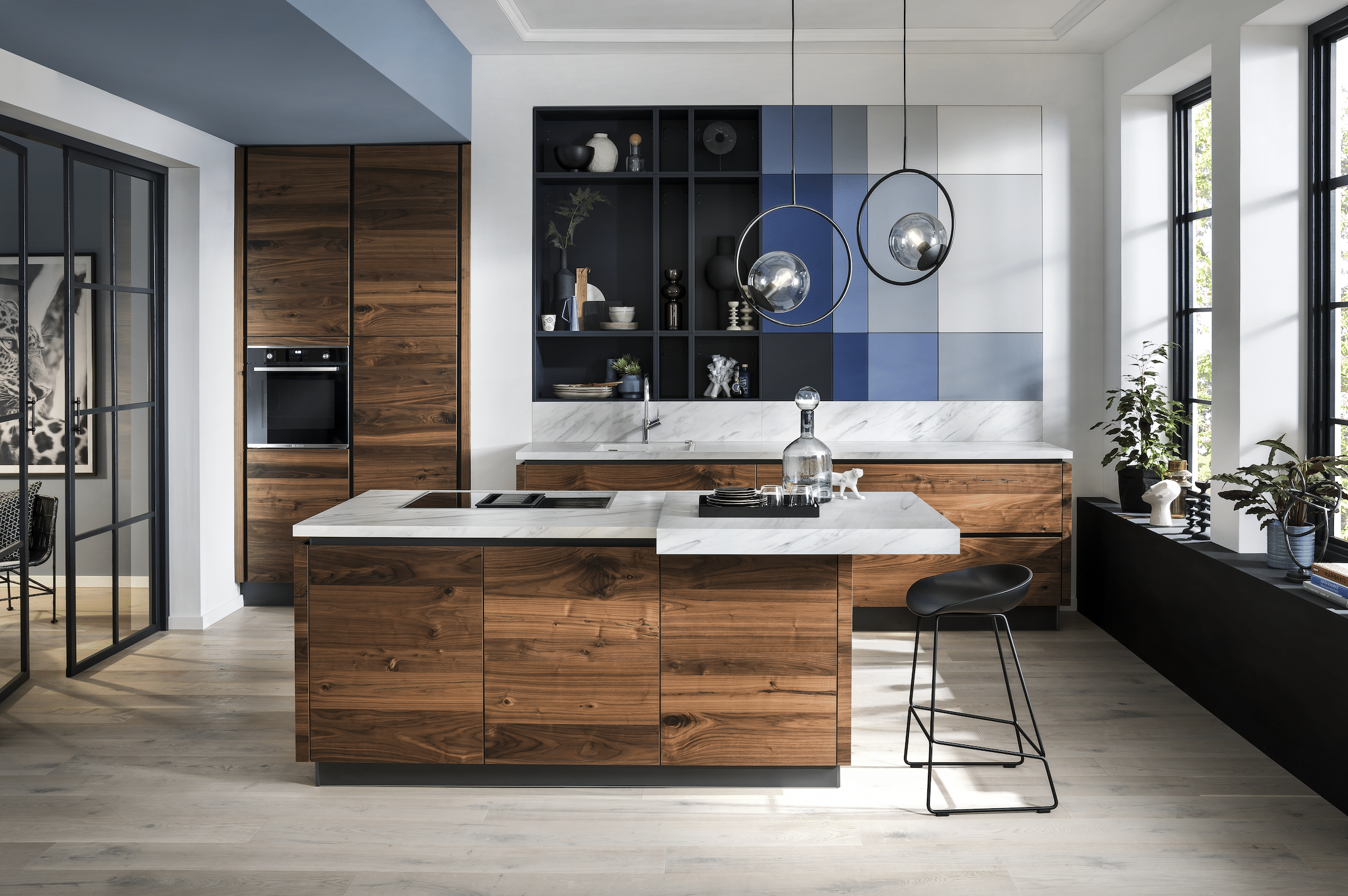 BÖHM MÖBEL zeigt eine Häcker Küche aus Holz, weißen Arbeitsplatten mit Marmorierung, blauen Elementen in der Wand und einem Barhocker.