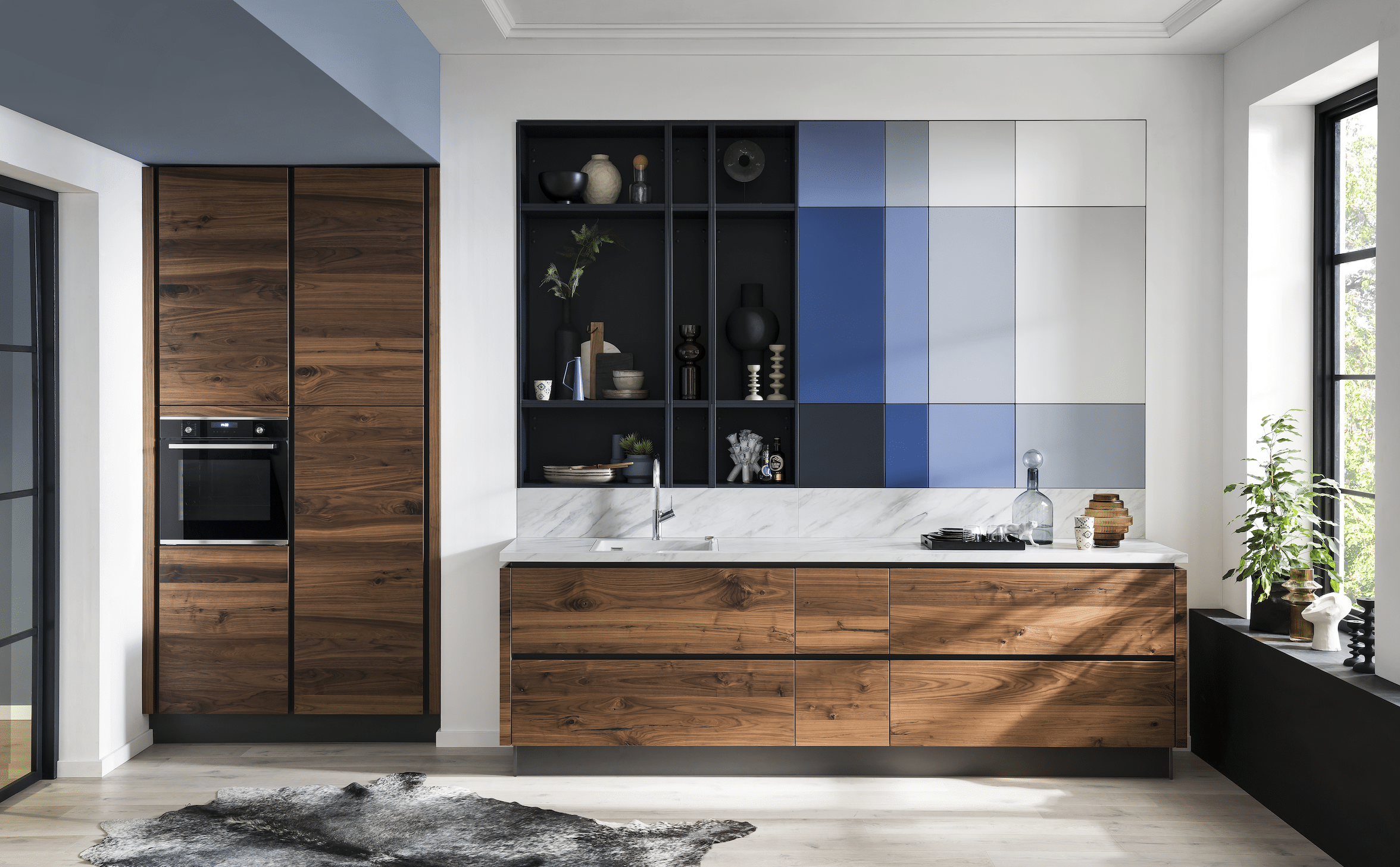 BÖHM MÖBEL zeigt eine Häcker Küche aus Holz, weißen Arbeitsplatten mit Marmorierung, blauen Elementen in der Wand und einem Barhocker.