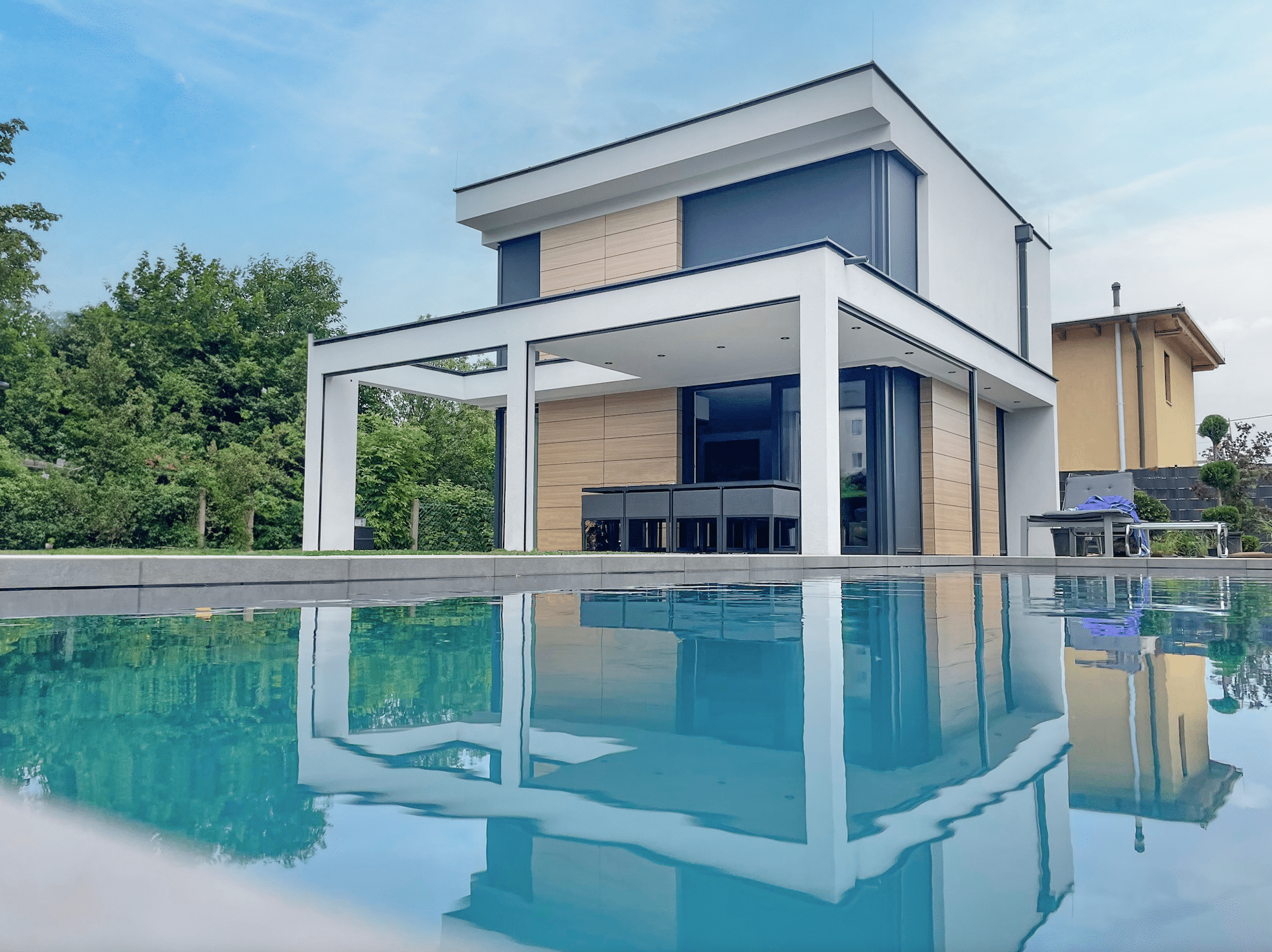 DIALOG Haus präsentiert ein modernes Einfamilienhaus mit Flachdach, weißer Putzfassade inkl. gemauerter Terrassenüberdachung mit Säulen und großem Pool davor.