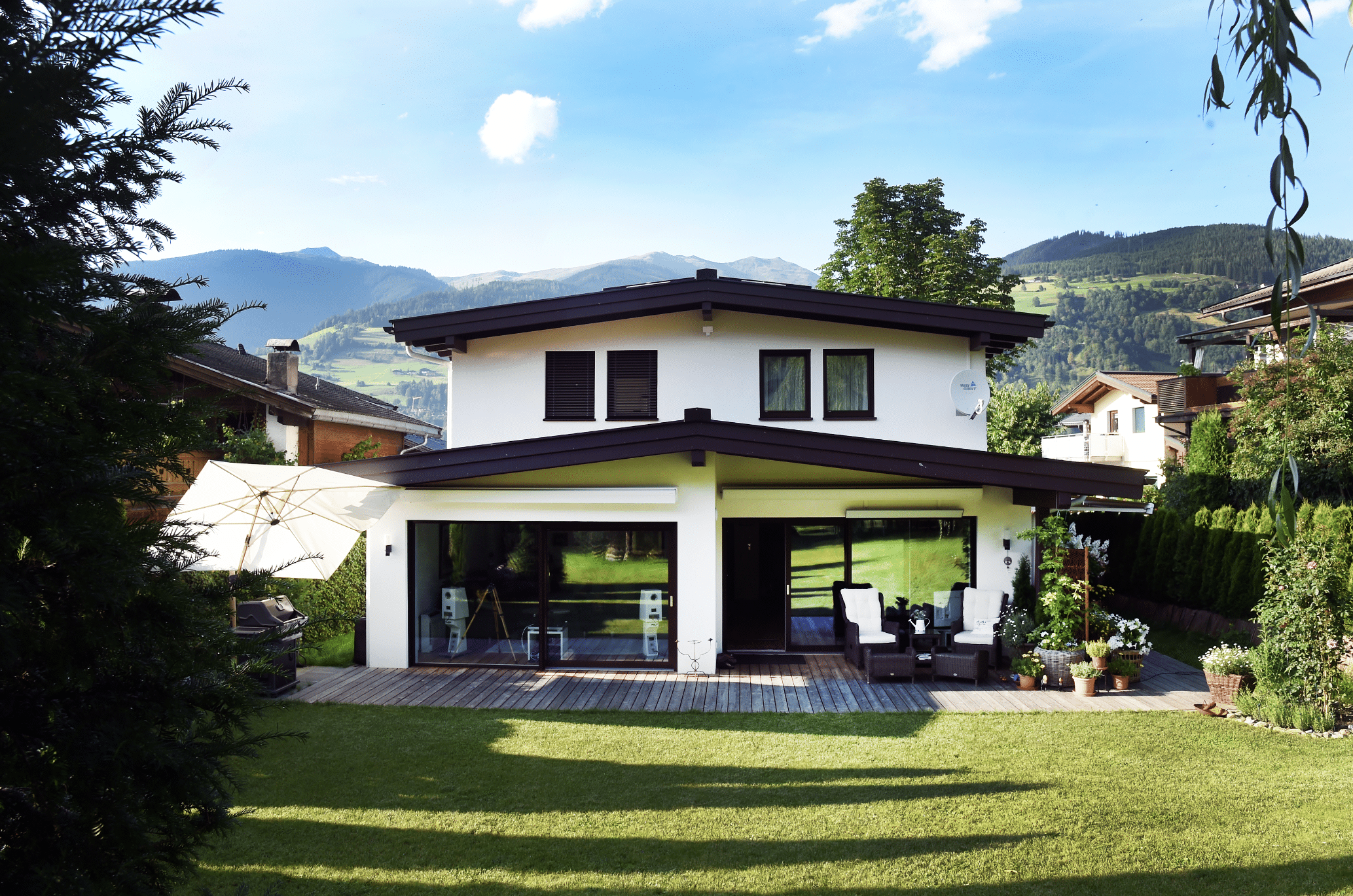 ERLER BAU GmbH zeigt ein weisses Einfamilienhaus mit 2 Stockwerken und einer überdachten Terrasse mit Holzboden und einer gemütlichen Sitzecke mit Rattanmöbeln und Glasschiebetüren mit Sicht auf den Wohnbereich.