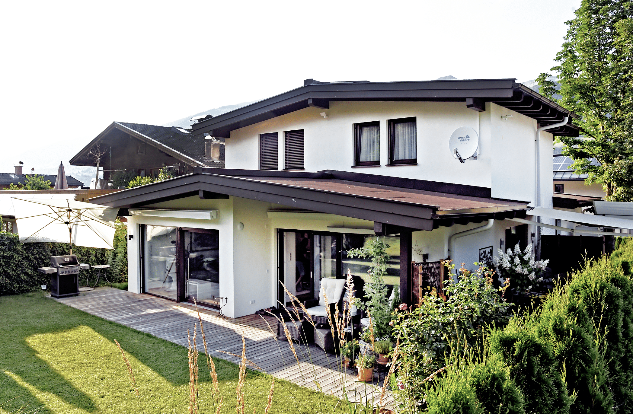 ERLER BAU GmbH zeigt ein weisses Einfamilienhaus mit 2 Stockwerken und einer überdachten Terrasse mit Holzboden und einer gemütlichen Sitzecke mit Rattanmöbeln, einem großen Sonnenschirm und einem Grill.