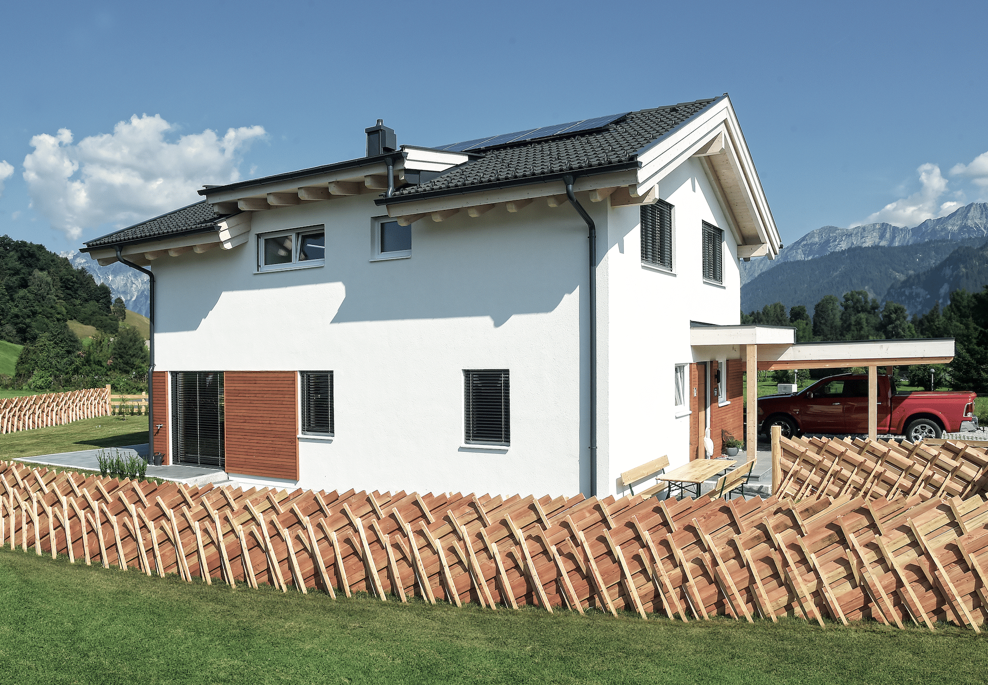 ERLER BAU GmbH zeigt ein weisses Einfamilienhaus mit Holzfassade, einem Carport und hellem Holzzaun vor dem gepflegten Garten mit gefliester Terrasse.
