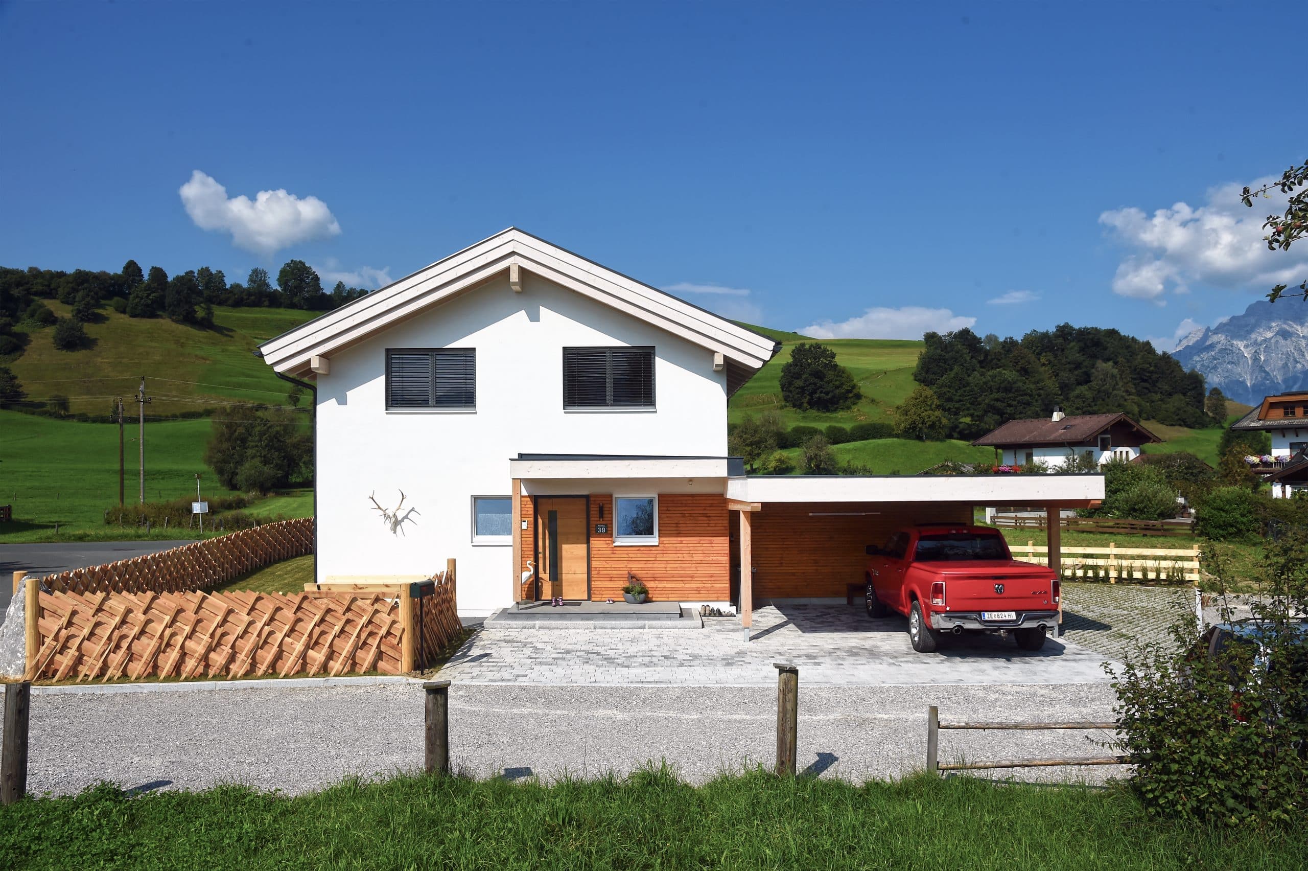 ERLER BAU GmbH zeigt ein weisses Einfamilienhaus mit Holzfassade und einem Carport aus Holz, mit dunklem Dach und einem Gartenzaun aus hellem Holz.