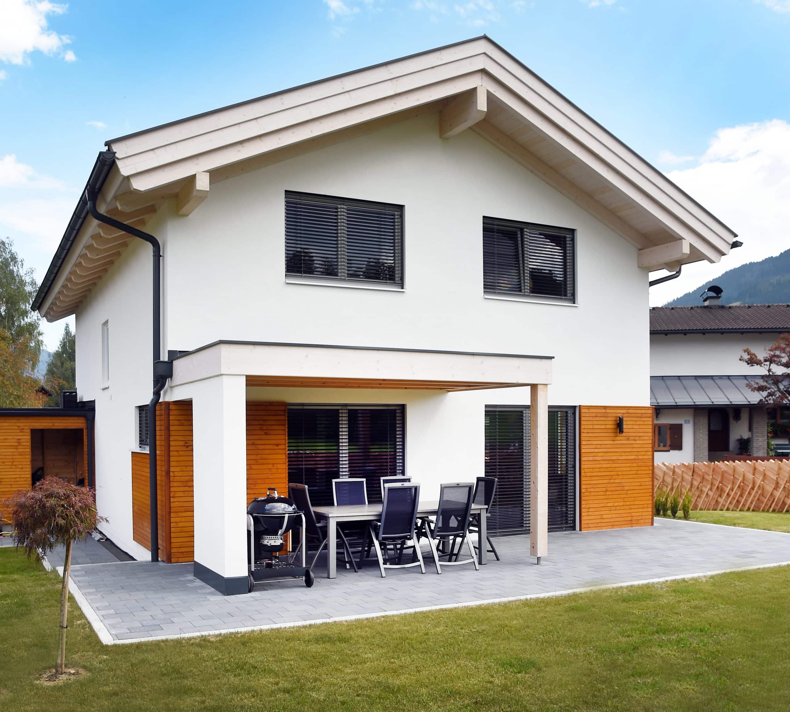 ERLER BAU GmbH zeigt ein weisses Einfamilienhaus mit Holzfassade und einer gefliesten Terrasse mit gemütlicher Sitzgruppe und schwarzem Griller.