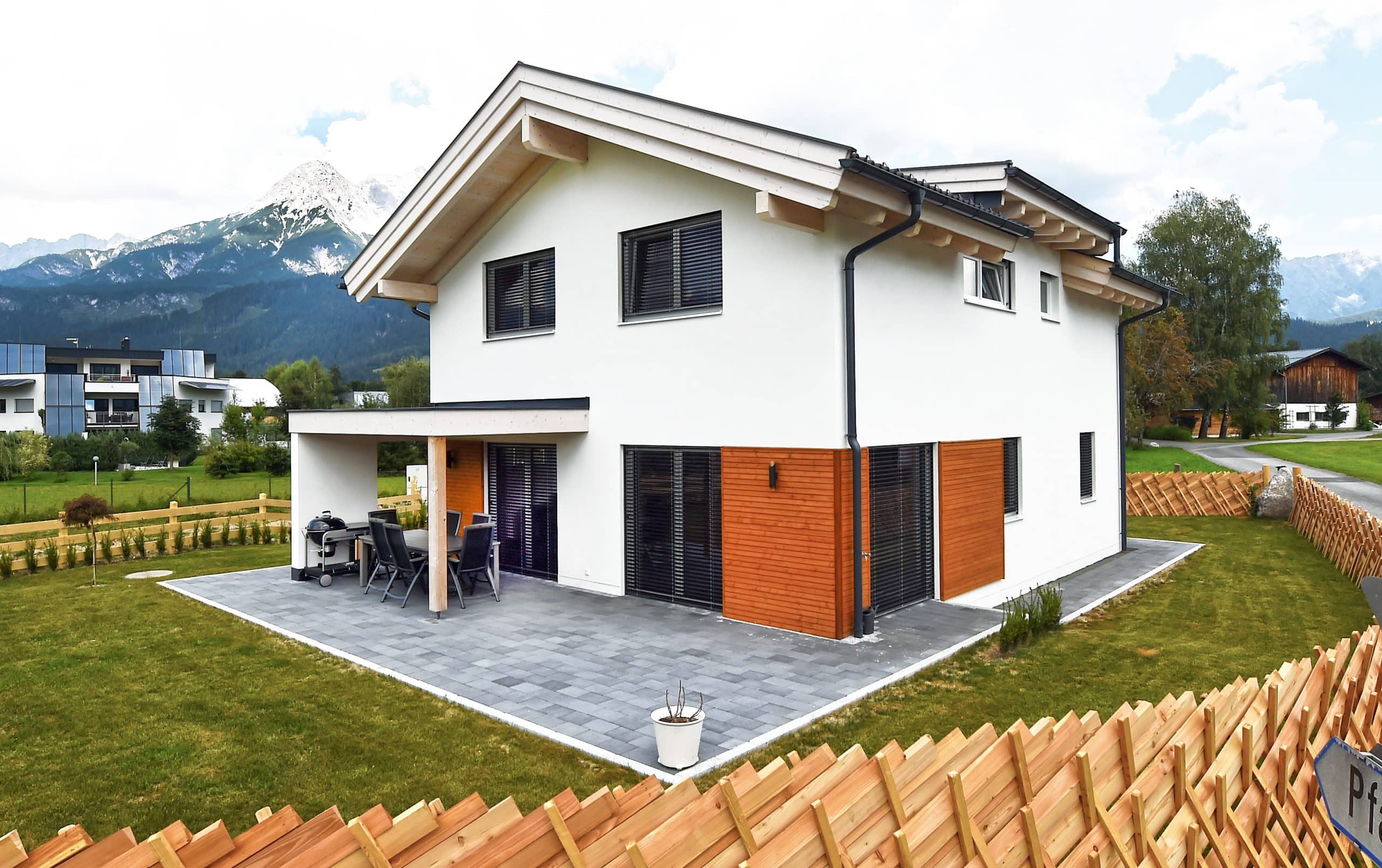 ERLER BAU GmbH zeigt ein weisses Einfamilienhaus mit Holzfassade und einer gefliesten Terrasse mit gemütlicher Sitzgruppe und einem hellen Holzzaun.