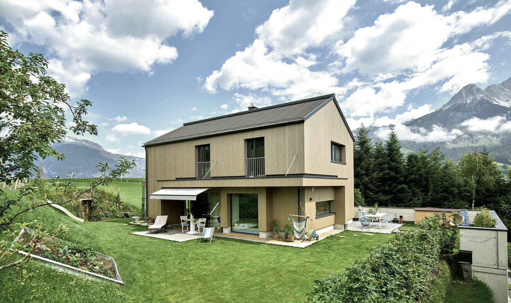 ERLER BAU GmbH zeigt ein modernes Einfamilienhaus aus Holz mit großen Fenstern und einer mit Markise überdachten Terrasse mit Fliesenboden und gemütlichen Möbeln im gepflegten großen Garten.