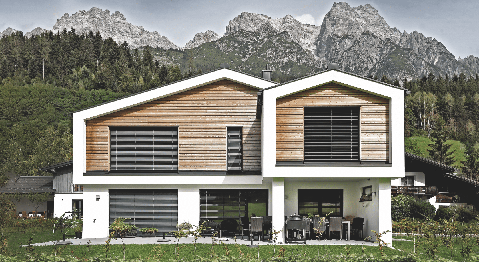 ERLER BAU GmbH zeigt ein modernes Einfamilienhaus mit Holzelementen, großen Fenstern an denen Rollläden montiert wurden und einer überdachten Terrasse mit einer Sitzgruppe.