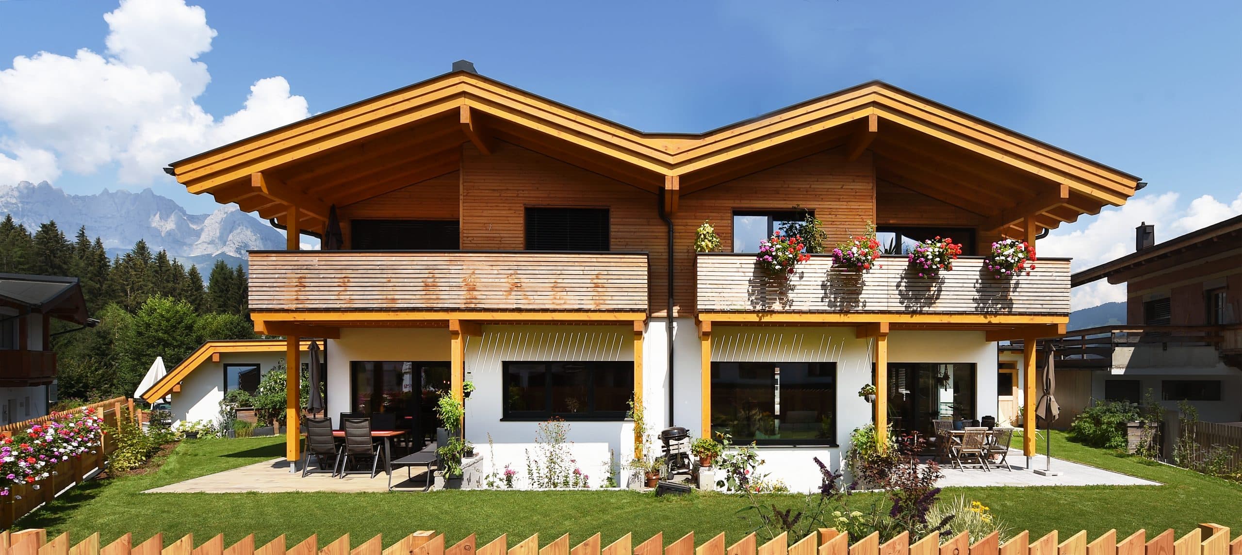 ERLER BAU GmbH zeigt ein schönes, rustikales Holzhaus mit großem Balkon und einer überdachten Terrasse mit gemütlichen Möbeln und vielen Pflanzen.