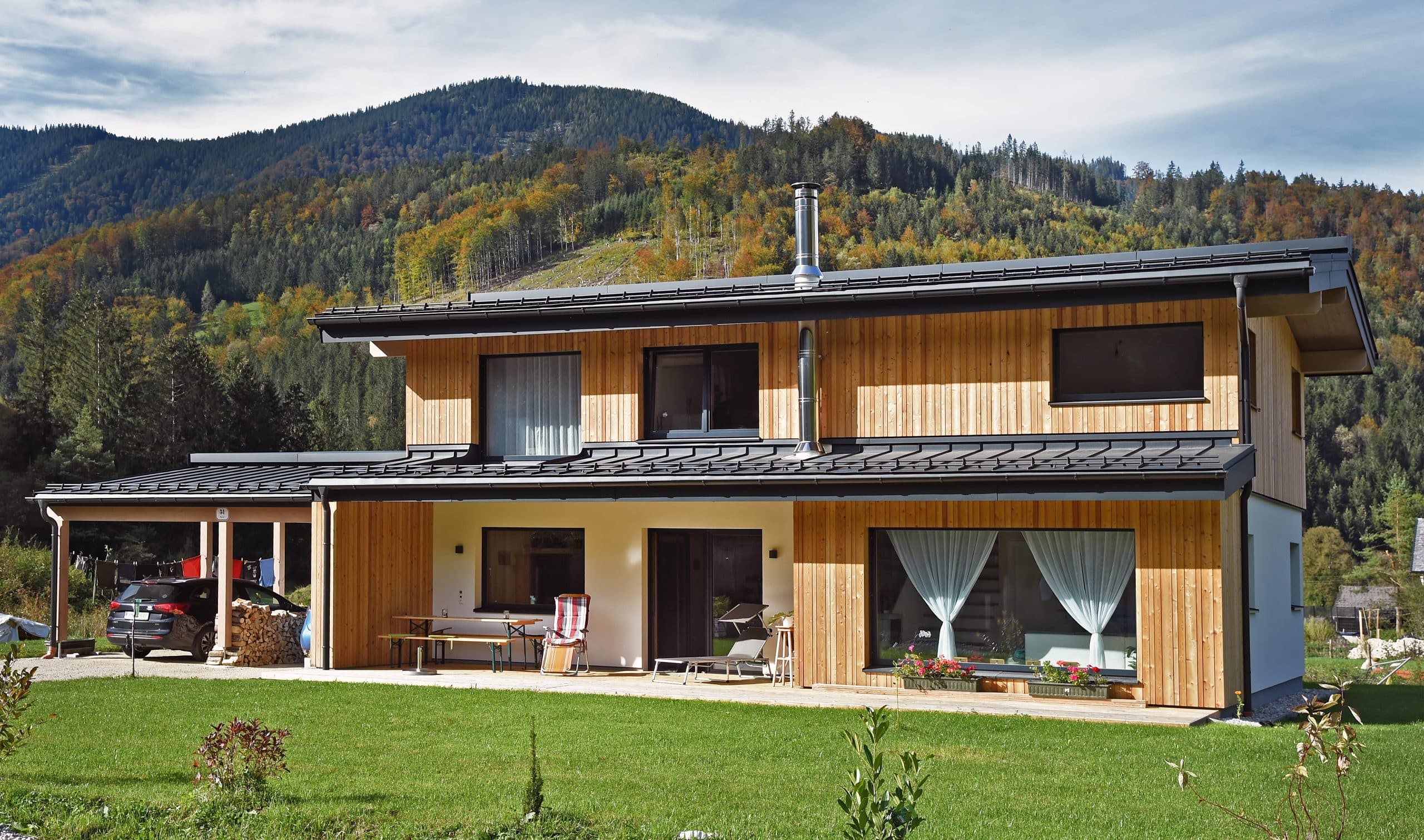 ERLER BAU GmbH zeigt ein großes Holzhaus im Grünen mit Carport und überdachter Terrasse mit einer Sitzecke.