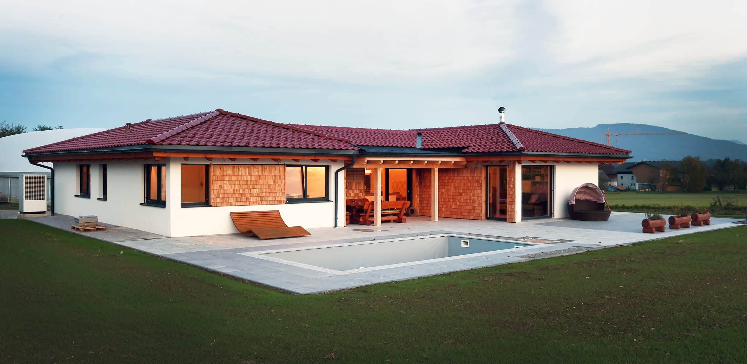 Ein schöner Bungalow mit rotem Dach, überdachter Terrasse mit einer Sitzgruppe aus Holz und großem Garten mit rechteckigem Pool von ERLER BAU GmbH.