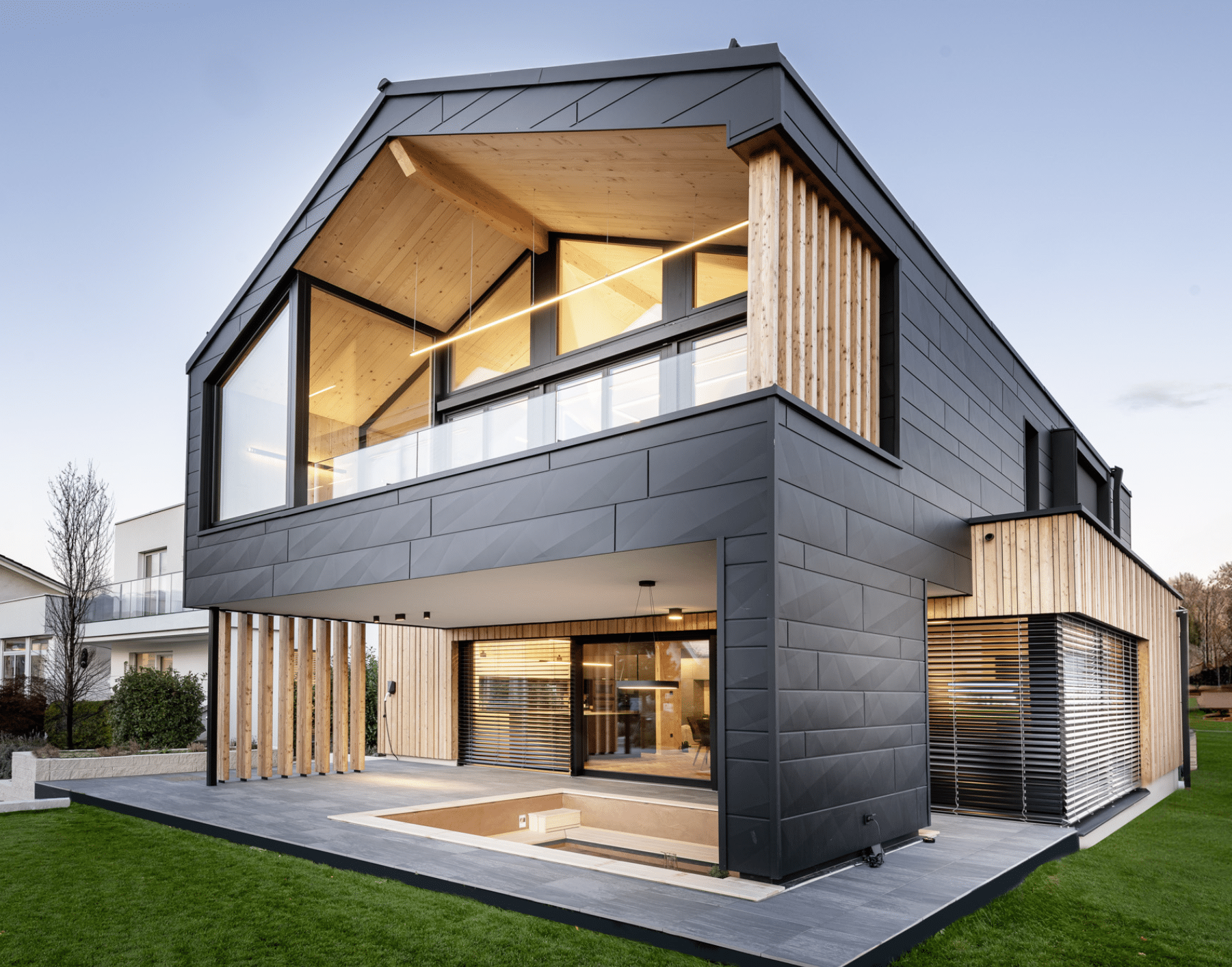 Erler Haus zeigt ein sehr modernes Holzriegelhaus mit gefliester Terrasse,dunkler Wandverkleidung, Balkon und eingebauter Sitzlounge inklusive Fernseher.