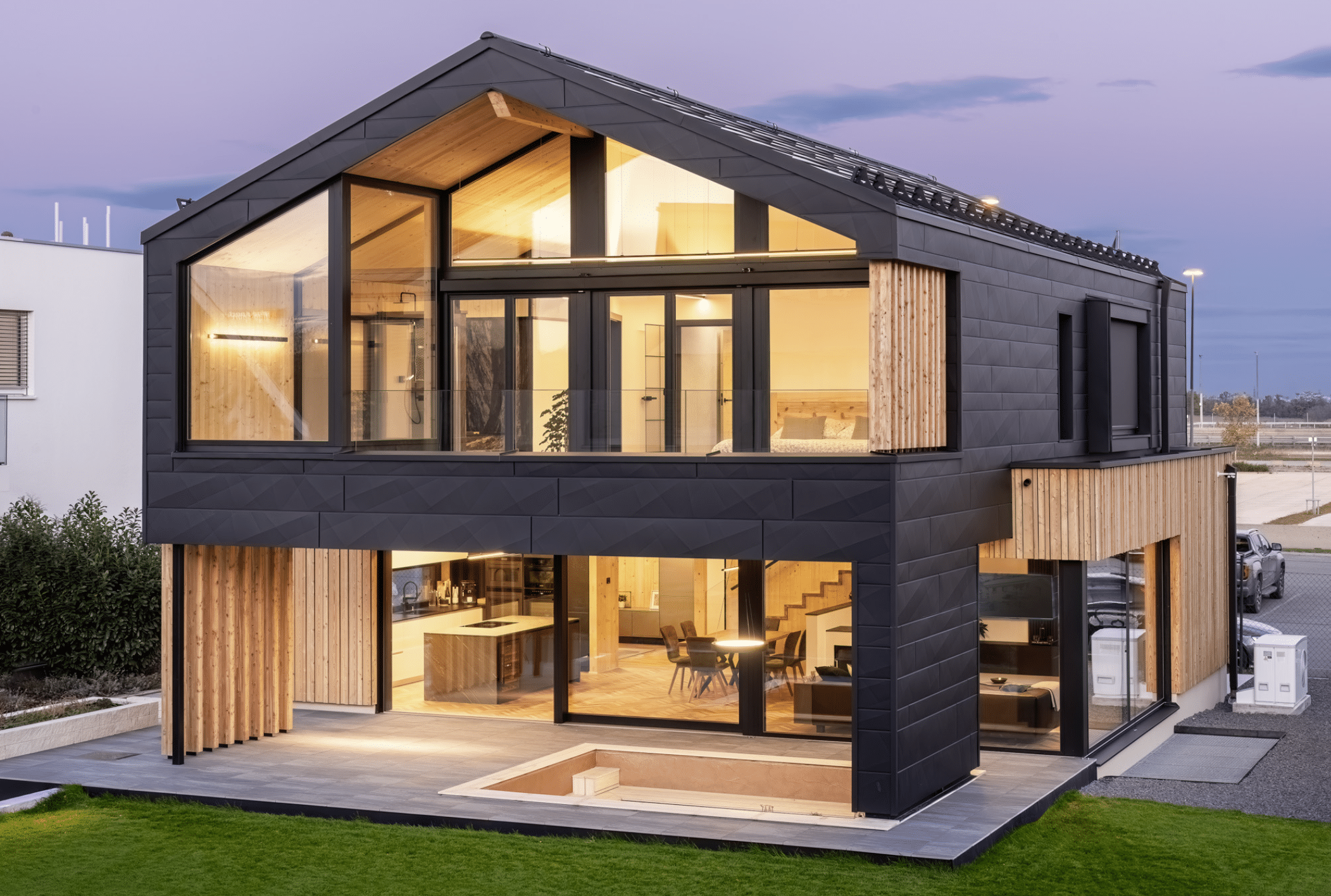Erler Haus zeigt ein sehr modernes Holzriegelhaus mit gefliester Terrasse,dunkler Wandverkleidung, Balkon und eingebauter Sitzlounge inklusive Fernseher bei Dämmerung.