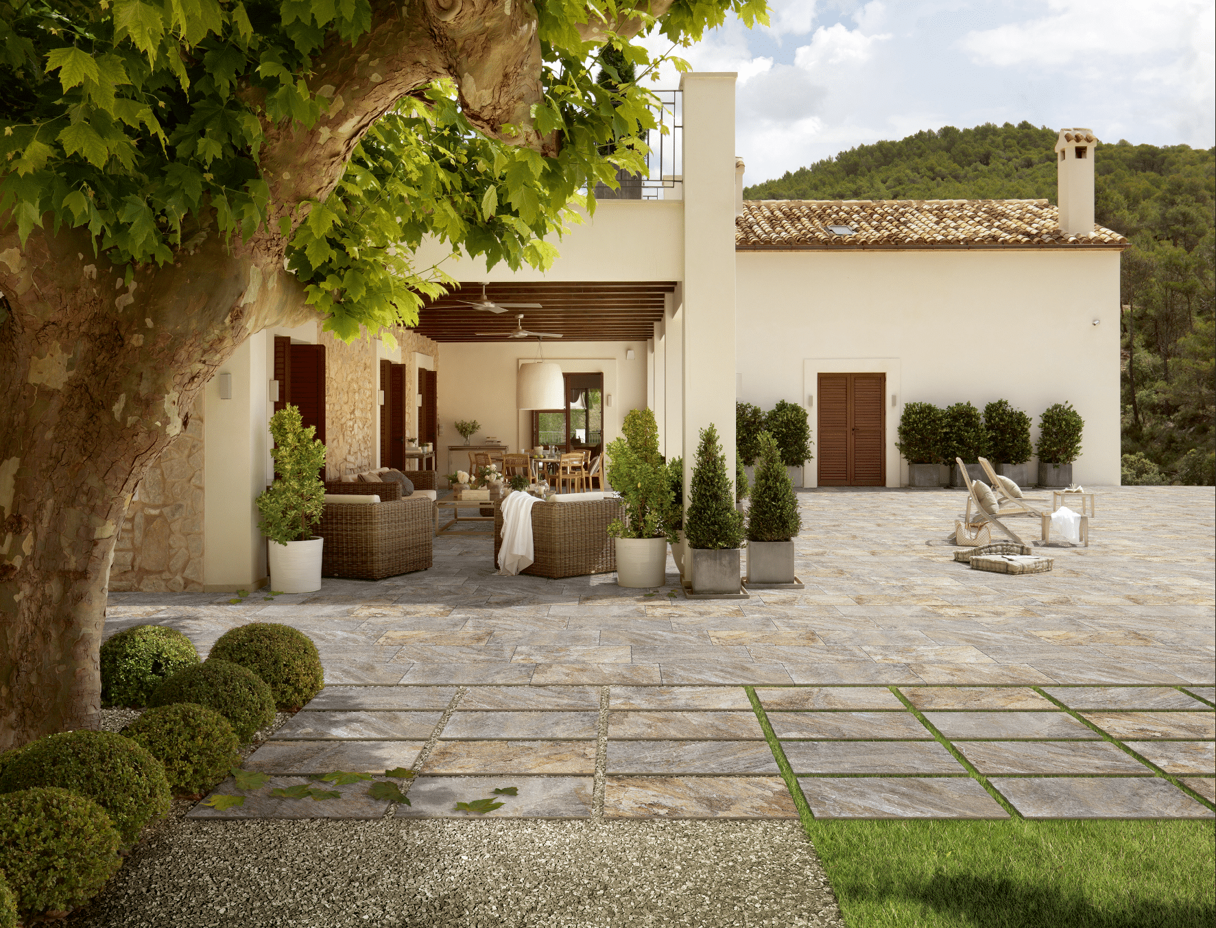 Mediterranes Landhaus samt Terrasse mit Steinzeug von Fliesendorf, ausgestattet mit Topfpflanzen, Liegestühlen und Rattanmöbeln für den Garten.