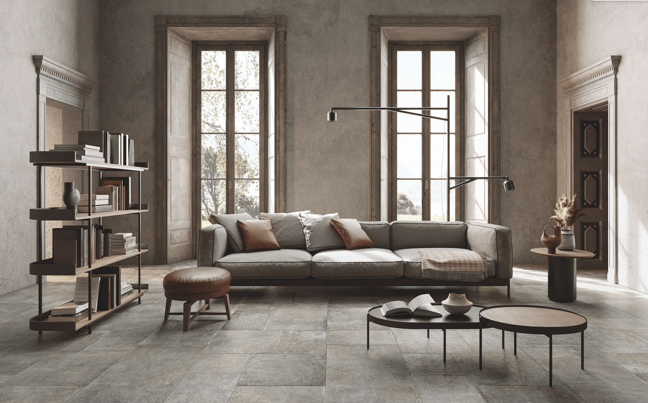 Fliesendorf zeigt ein Wohnzimmer mit grau gefliestem Boden, einer grauen, gepolsterten Couch mit braunem Lederhocker und zwei wandhohe Fenster mit Holzrahmen.