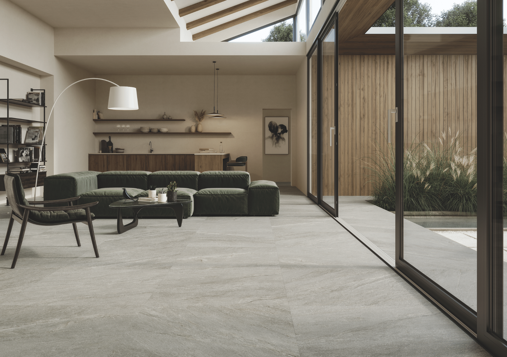 Fliesendorf zeigt ein modernes Wohnzimmer mit grauem Fliesenboden und großer Sitzlandschaft in dunklem grün und einer Glasschiebetür mit Zugang zur Terrasse mit vielen Pflanzen.