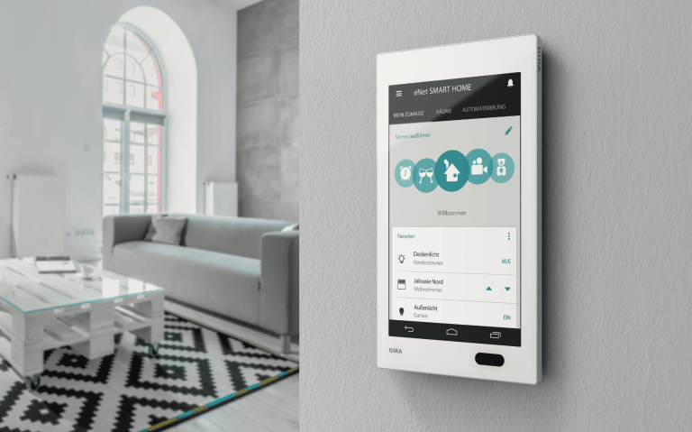 Gira zeigt eine weisse Smart Home Steuerung, mit großem Display, an der Wand zu einem Wohnzimmer mit grauer Couch, einem Palettentisch und einem Rundbogenfenster.