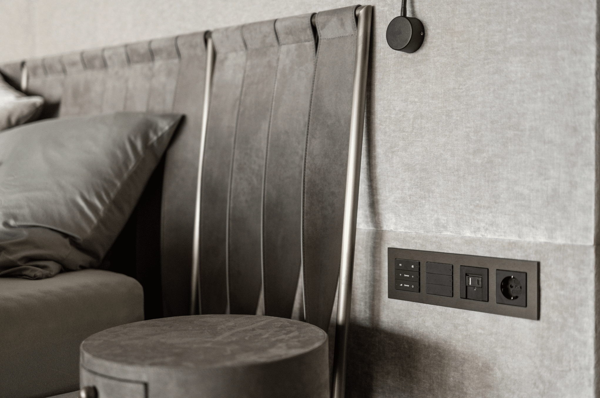 Gira zeigt den Kopfteil eines Doppelbetts mit einer modernen Steckerleiste in Metall-Optik und schwarzen Tasten und Steckdosen mit USB an der Wand daneben.