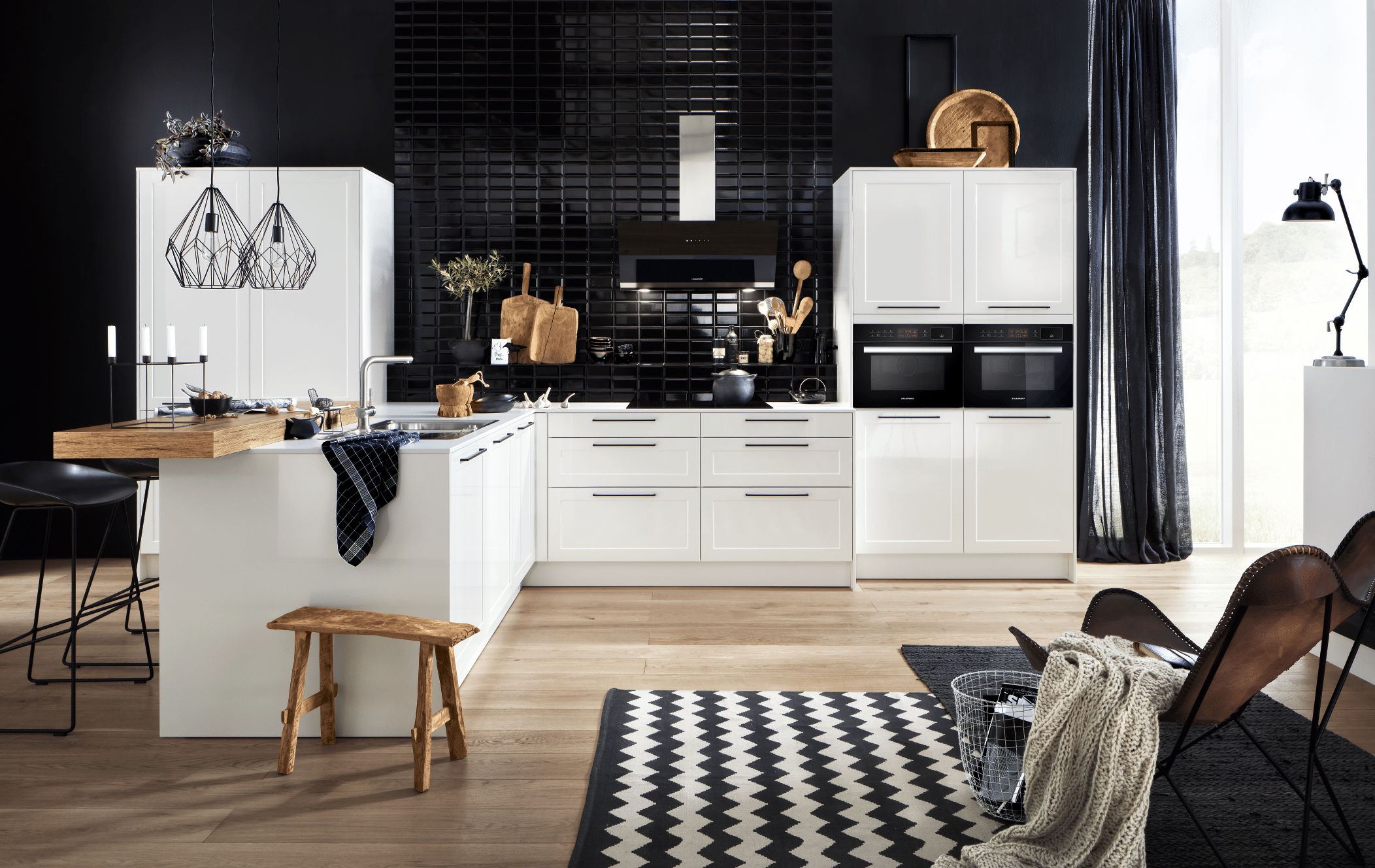 Offene Wohnküche mit Holzboden und Sitzgelegenheit sowie weißen Fronten und schwarzem Fliesenspiegel von Häcker Küchen, erhältlich im WOHNHAUS Grill & Ronacher.