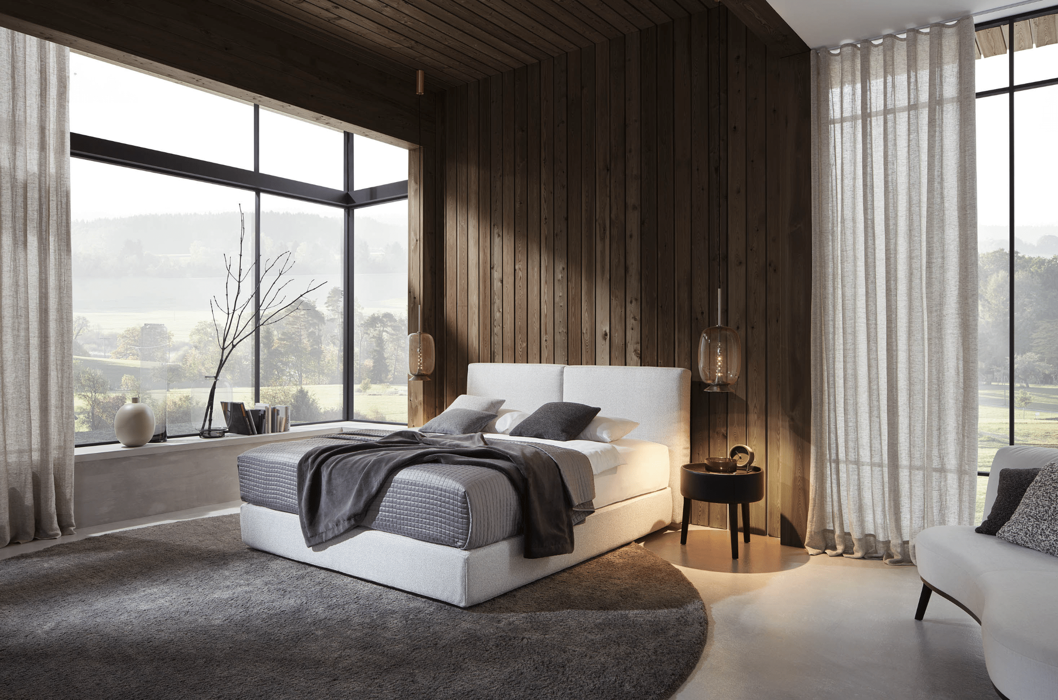 Grill & Ronacher zeigt ein lichtdurchflutetes Schlafzimmer mit weißem Doppelbett, rundem, grauem Teppich, Holzverbau an der Wand und hohen Fenstern von Bielefelder Werkstätten.
