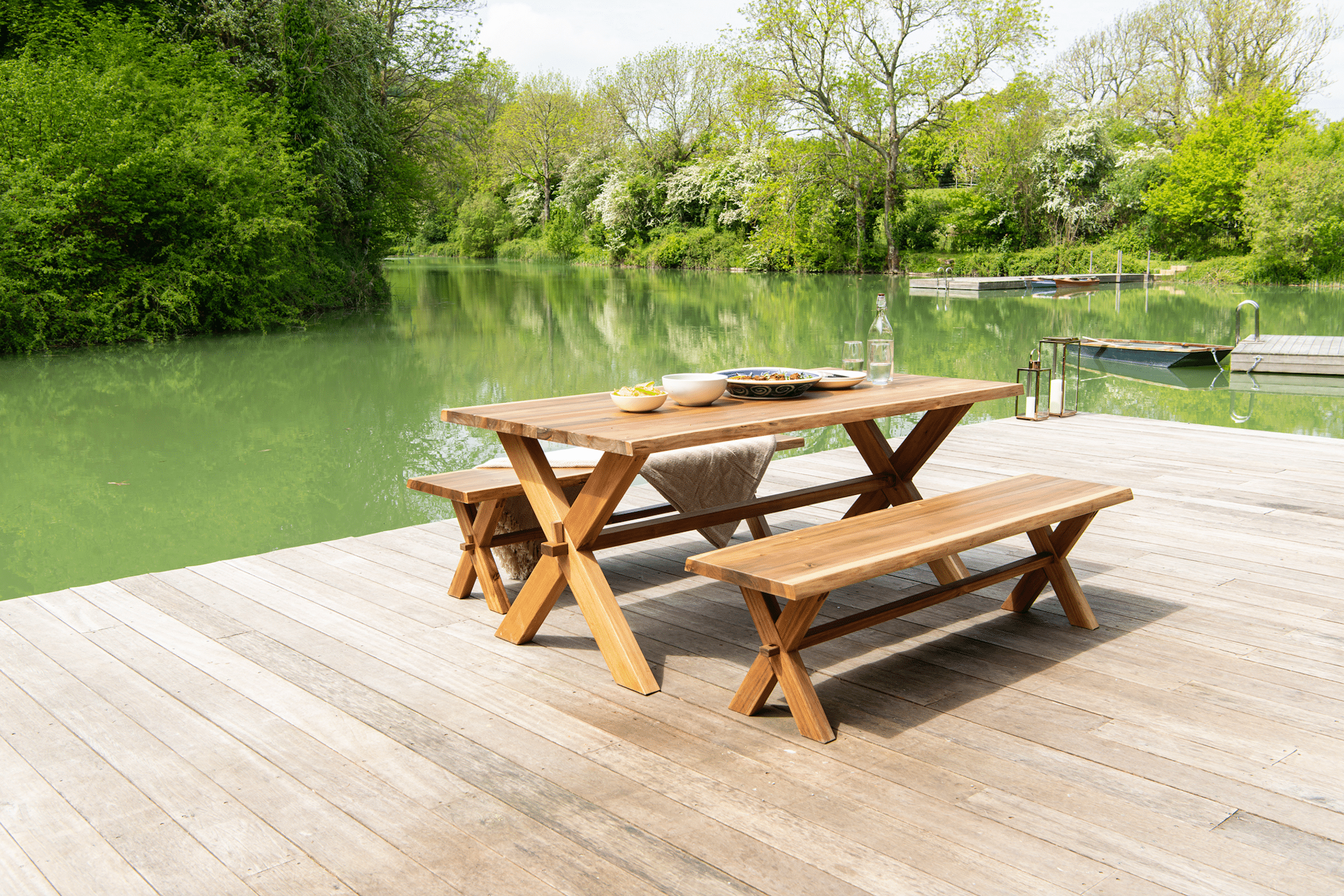 WOHNHAUS Grill & Ronacher zeigt eine gemütliche Sitzgruppe aus Holz mit einem großen Tisch und zwei Sitzbänken auf einem Holzboden mit Zugang zu einem See von Alexander Rose.