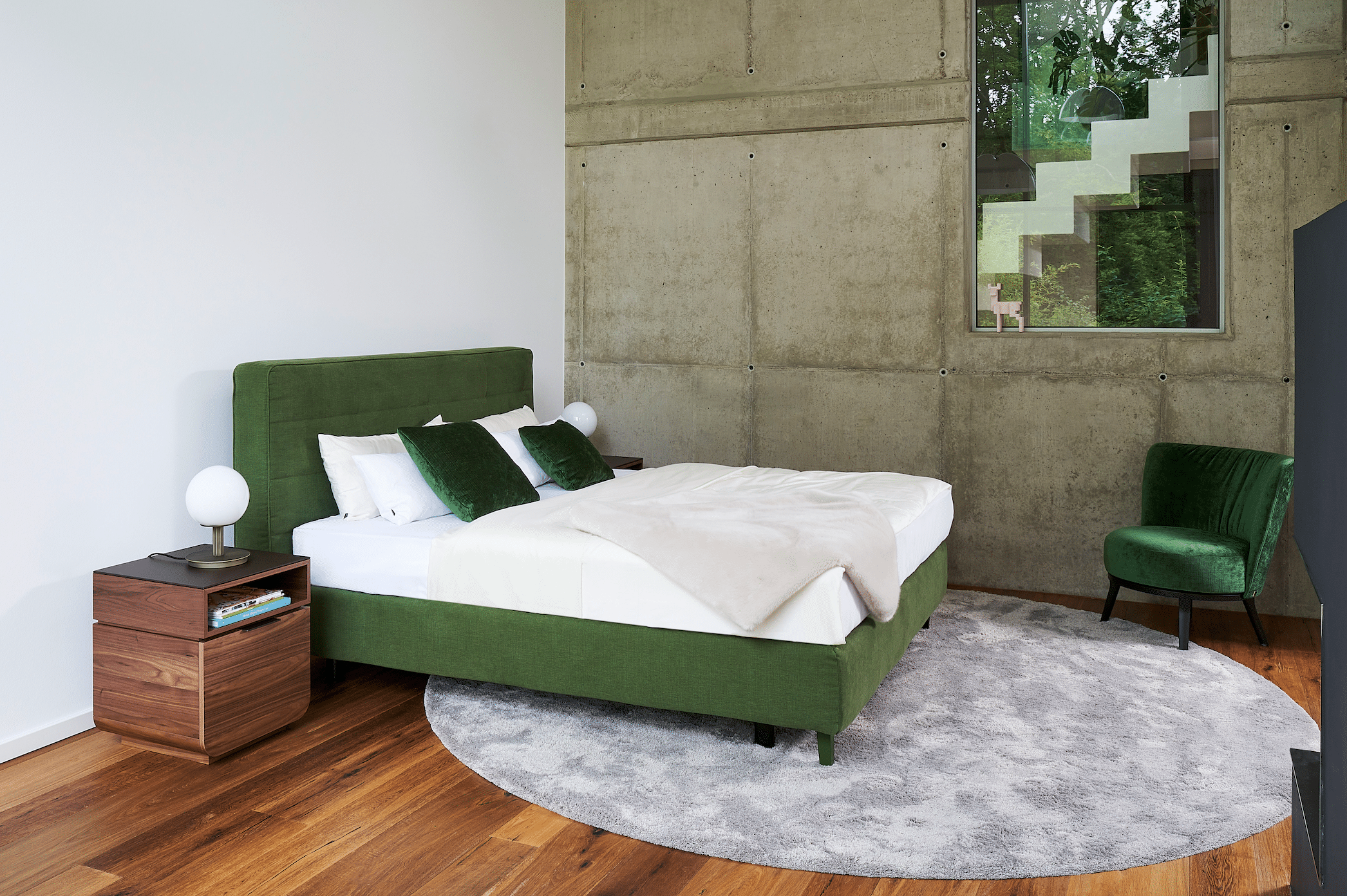 WOHNHAUS Grill & Ronacher zeigt ein Doppelbett in grün mit einem Nachtkästchen aus Holz und Ohrensessel aus Samt neben einer Wand im Industrial Look von Bielefelder Werkstätten.