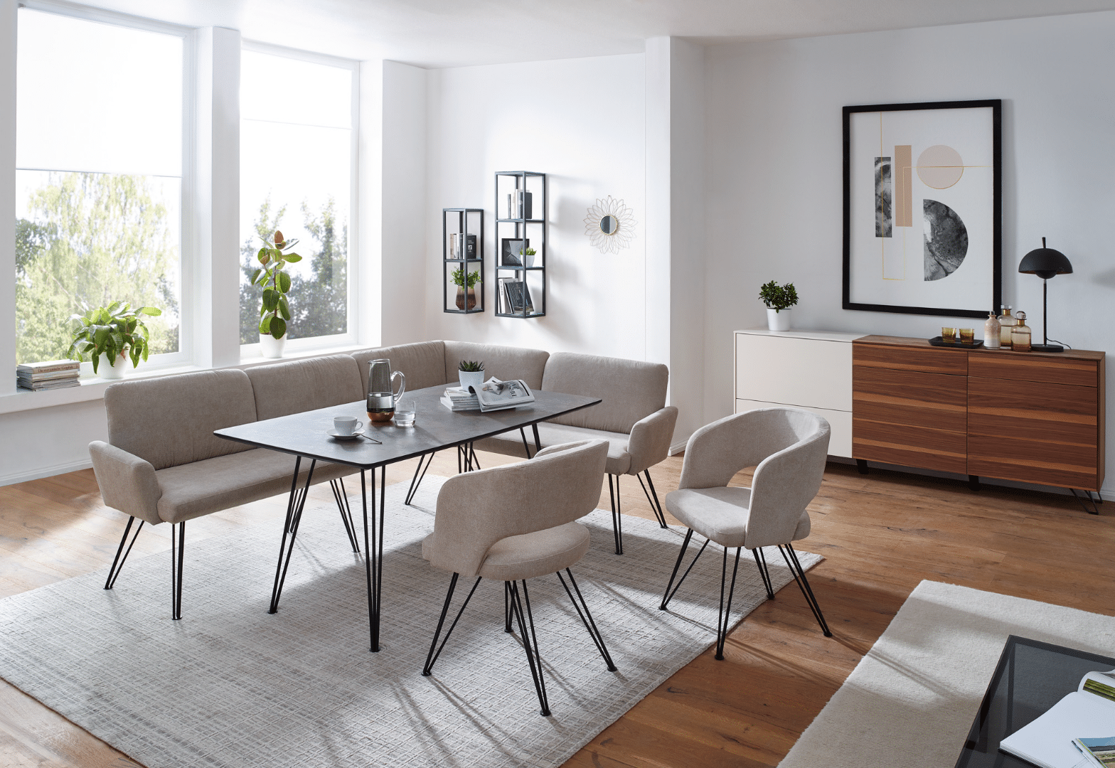 WOHNHAUS Grill & Ronacher zeigt helles Esszimmer mit Tisch, Stühle und Ecksitzbank mit beigem Stoff und Metallbeinen, dahinter eine Sideboard mit weißer und Holzfront und zwei hochformatige schmale Metallregale von Schösswender Möbel.