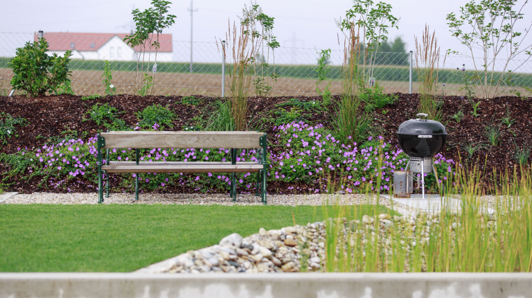 Grillplatz mit zwei Bänkchen in einem Garten mit lila Blümchen und Gartenzaun von Hablesreiter.