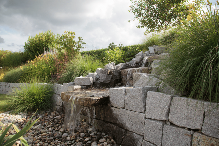 Hablesreiter zeigt hier eine Steinmauer mit integriertem Wasserfall der von oben herunter rinnt, mit viel Bepflanzung und verschiedenem Gras.