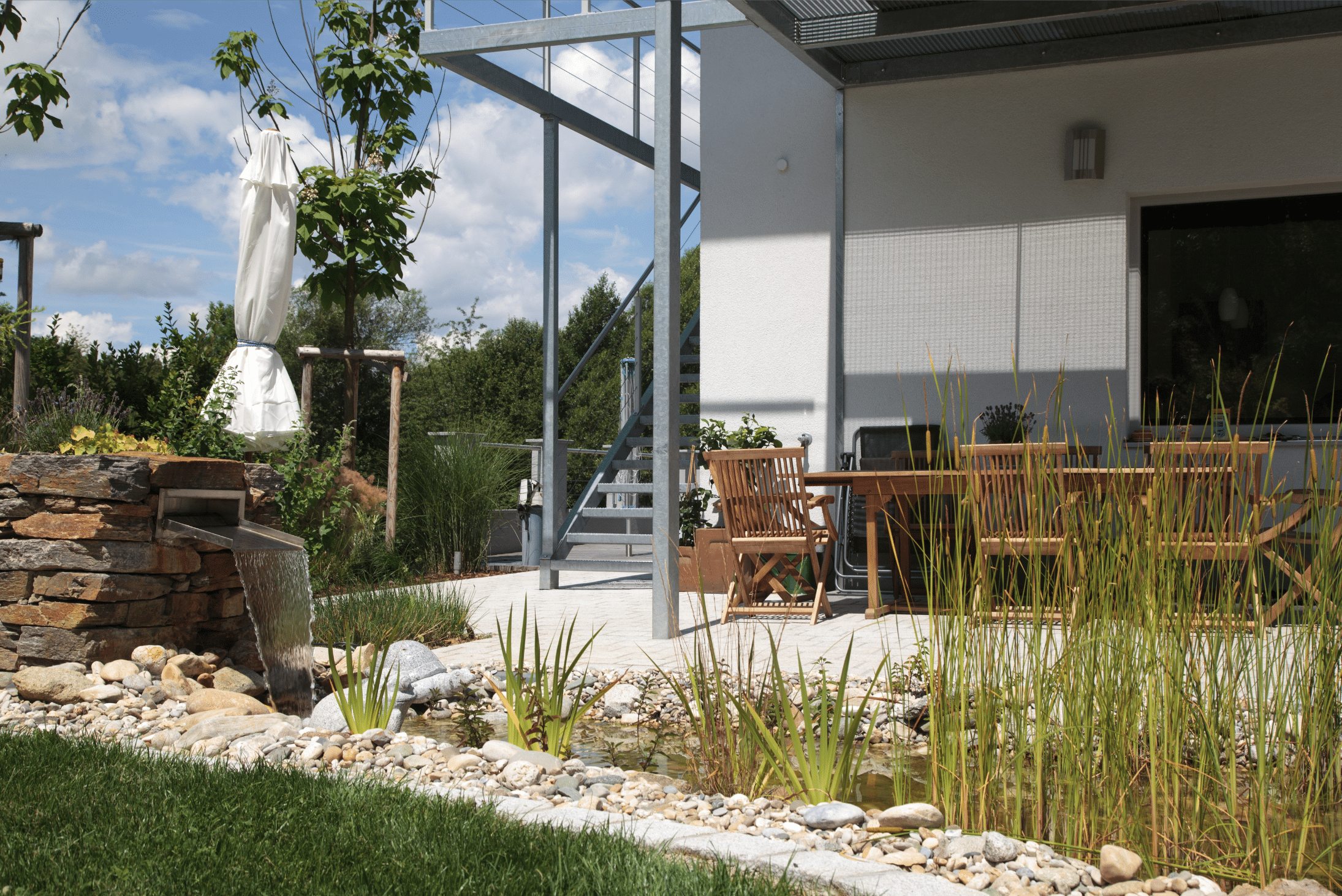 Hablesreiter zeigt eine überdachte Terrasse mit Gartenmöbel, einem kleinen Teich davor und ein integriertes Wasserspiel.