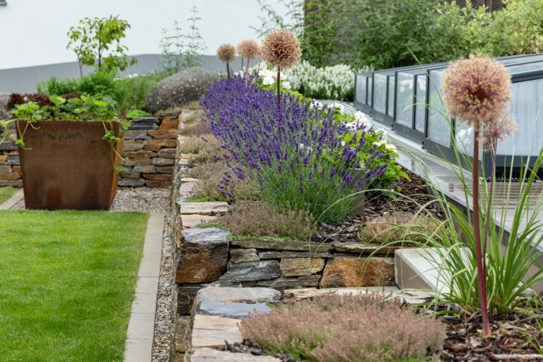 Beet mit Steinmauer, großer Blumentopf aus Eisen in rostiger Optik und Rasenfläche von Gartengestaltung Hablesreiter.