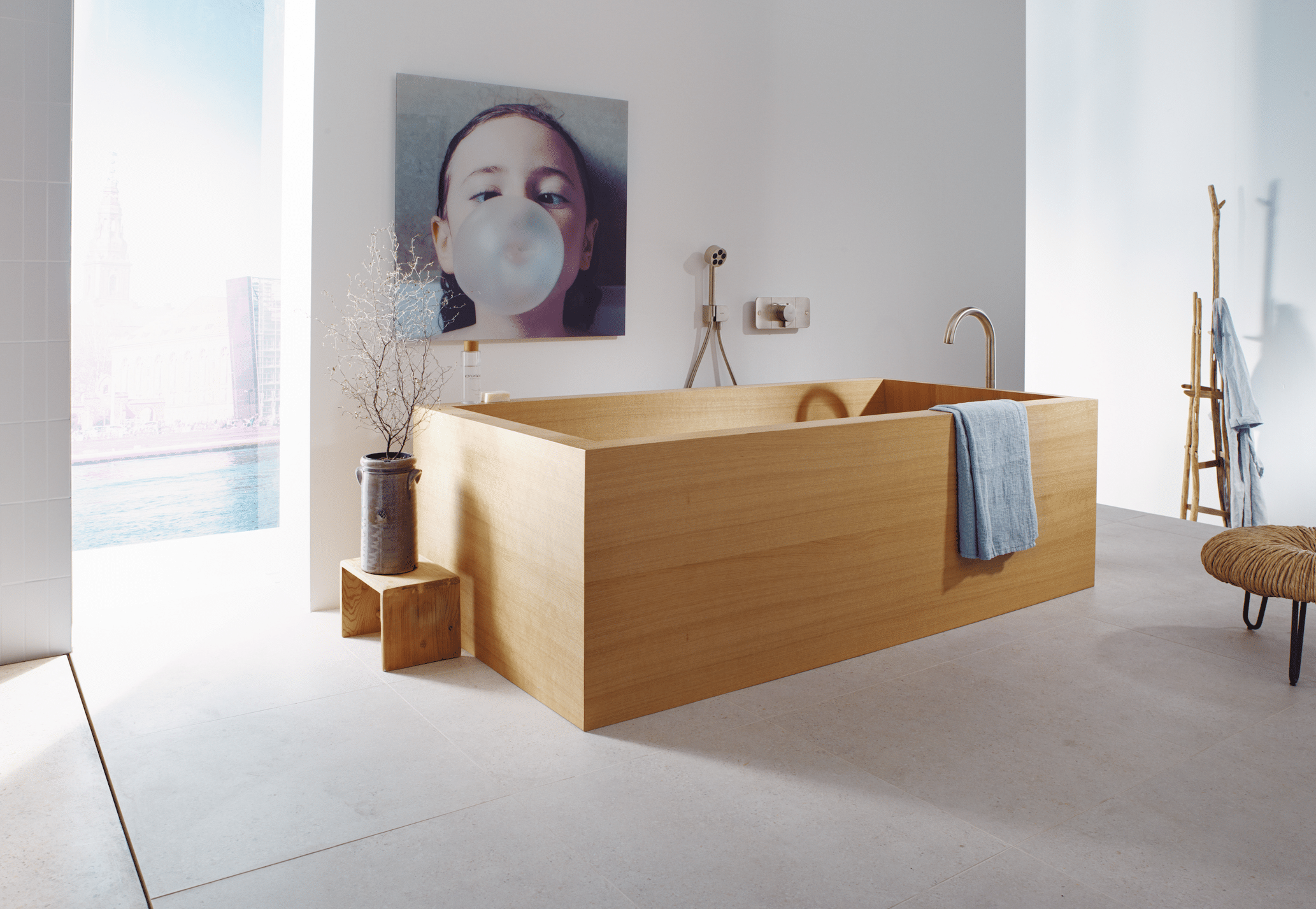 Hansgrohe präsentiert eine Badewanne in heller Holzoptik mit rose-goldener Armatur und einem Bild von einem Mädchen mit einer großen Kaugummiblase.