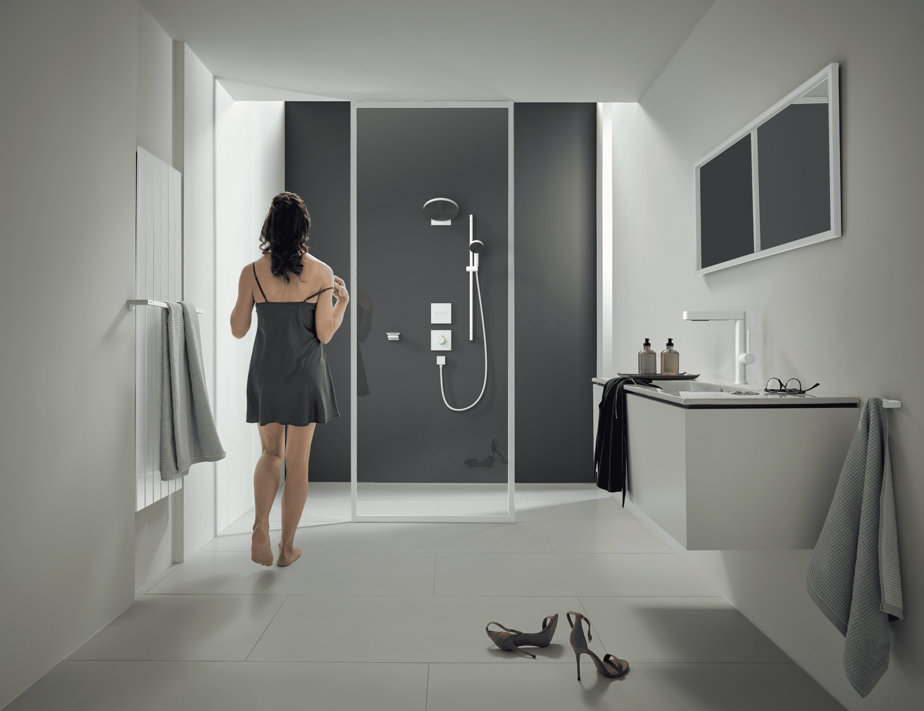 Hansgrohe präsentiert ein weißes Badezimmer mit grauer Duschrückwand, einer Glaswand und einer Dame die gerade in die Dusche spaziert.