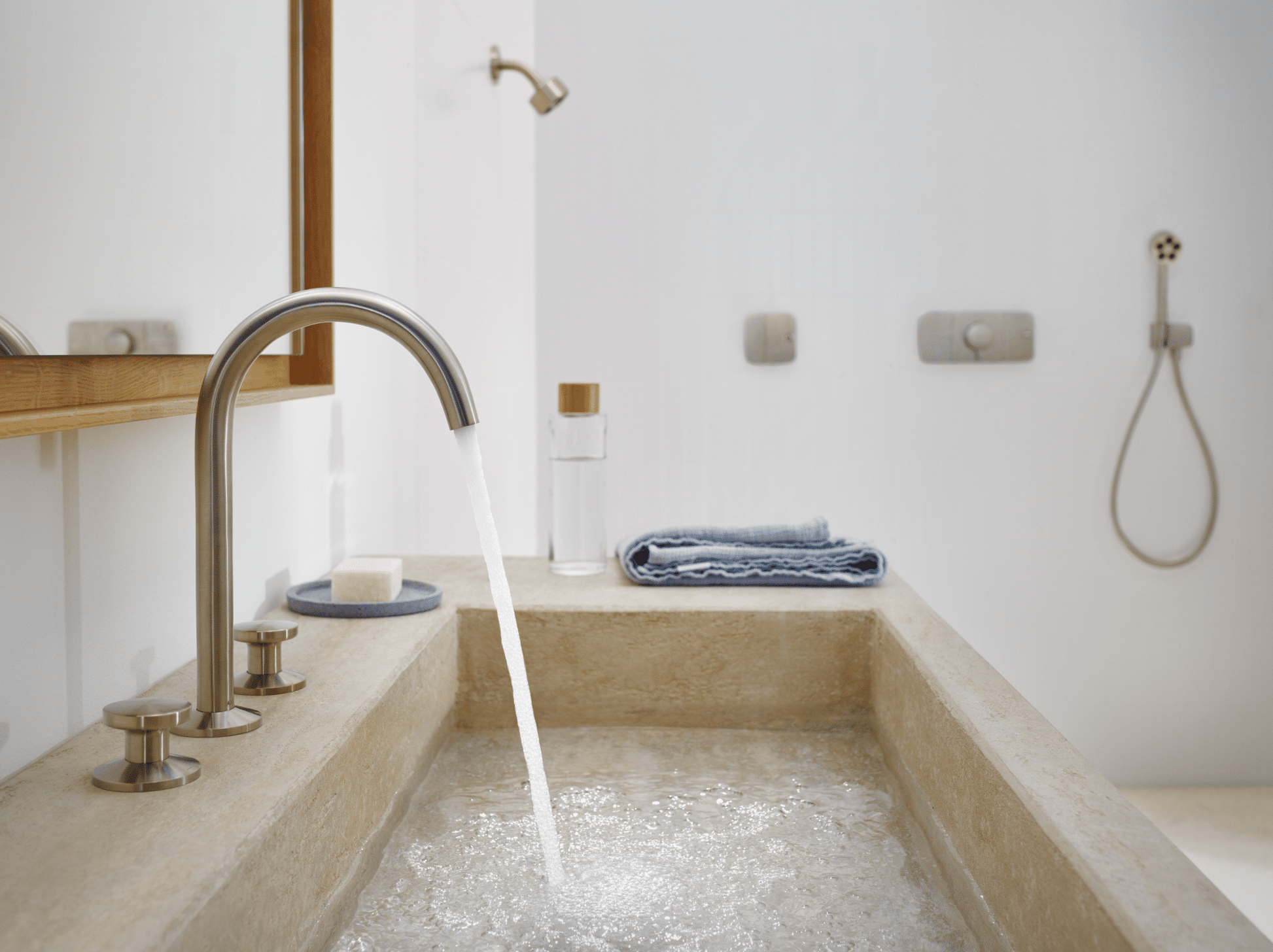 Hansgrohe zeigt ein Doppelwaschbecken in Kalksteinoptik mit goldenen Wasserhähnen, einem rechteckigen Spiegel und Walk-In Dusche.