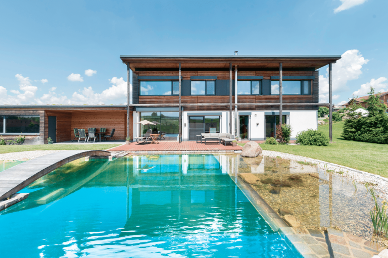 HARTL HAUS Holzindustrie zeigt ein modernes Fertighaus mit naturbelassener Holzfassade mit großem Schwimmteich und Terrasse.