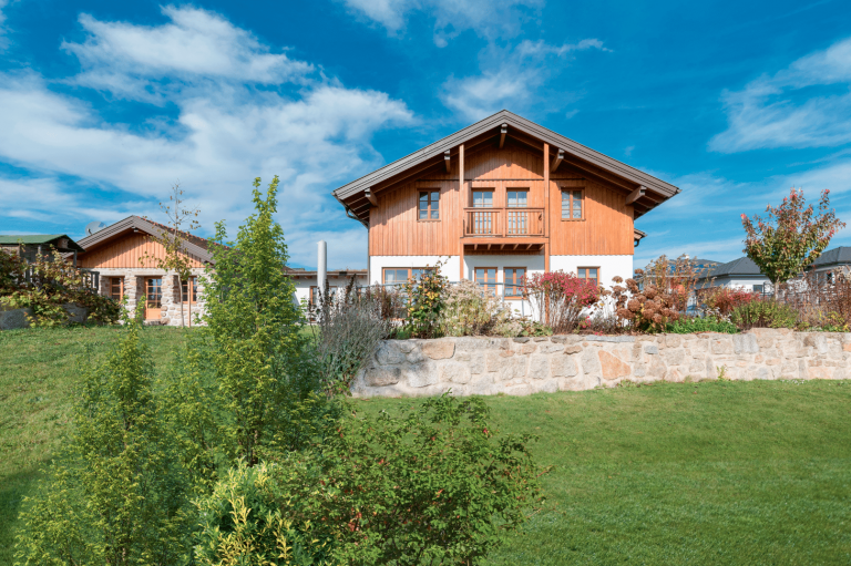 HARTL HAUS Holzindustrie zeigt ein Fertighaus im ländlichen Stil mit Satteldach, teilweise Holzfassade sowie Gartenanlage mit Natursteinmauer.