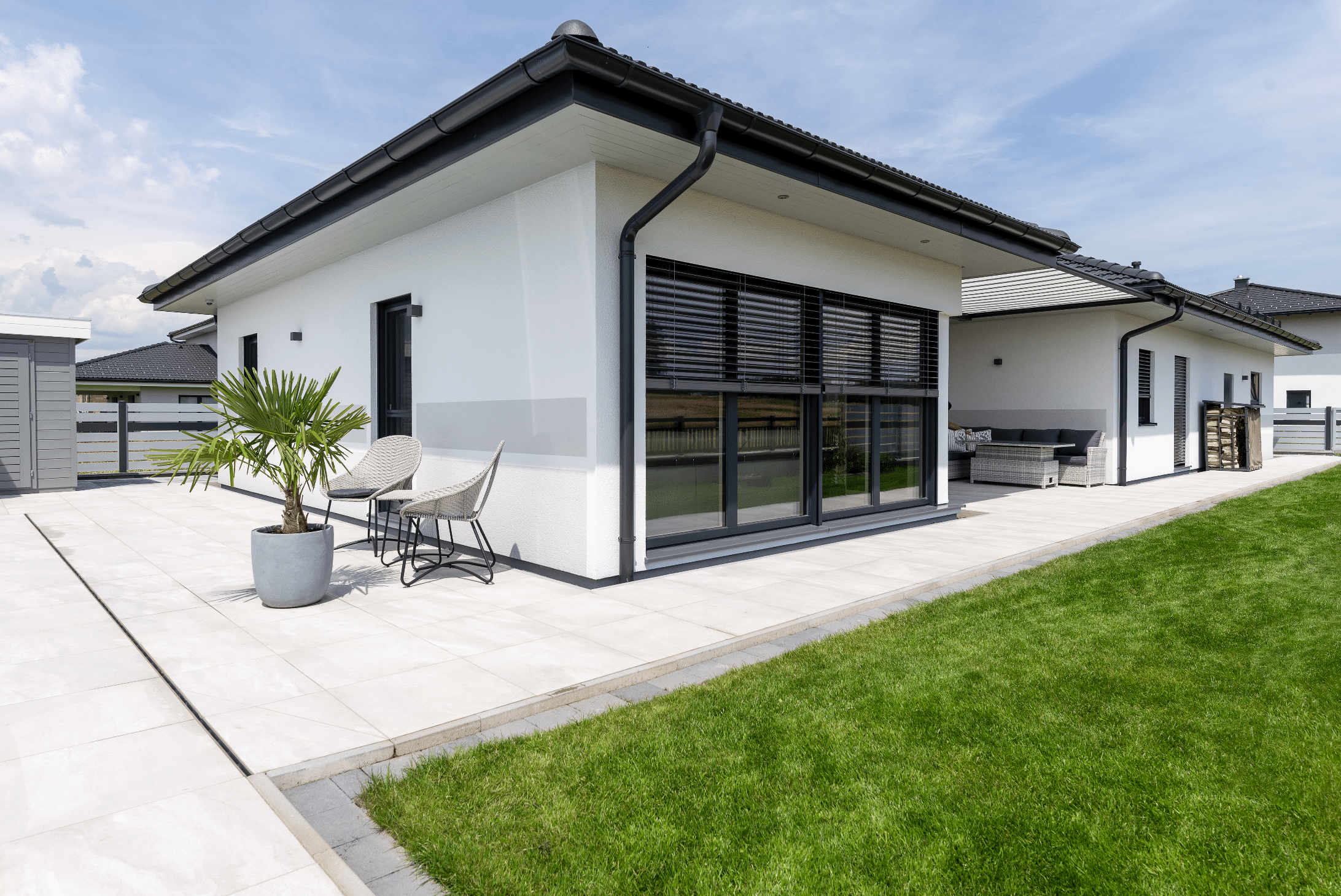 Weißer Bungalow von Hartl Haus, großer Terrassentür aus Glas mit dunklen-Rollläden und Gartenmöbeln.