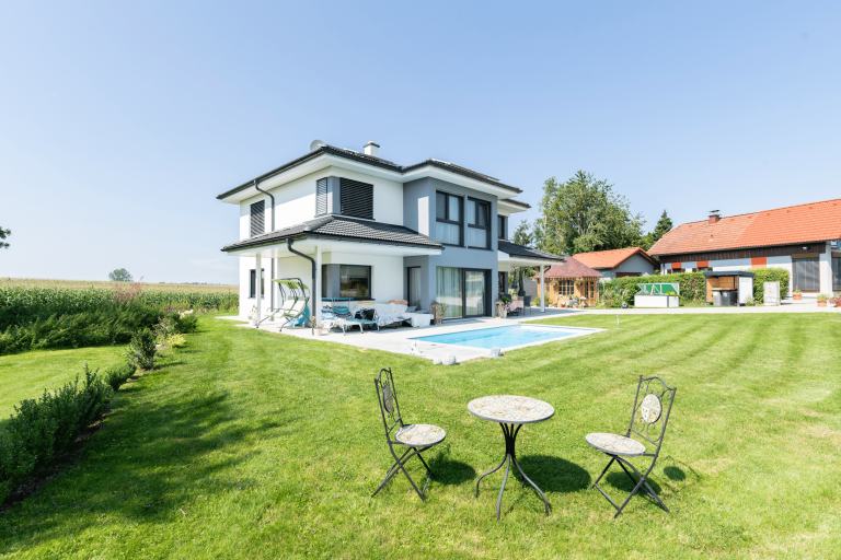 Weiß-graues eingeschößiges Haus von Hartlhaus mit großen Fenstern und dunklen Fensterrahmen großem Garten, kleinem Pool, Gartenhütte, Terrasse mit Liegestühlen.