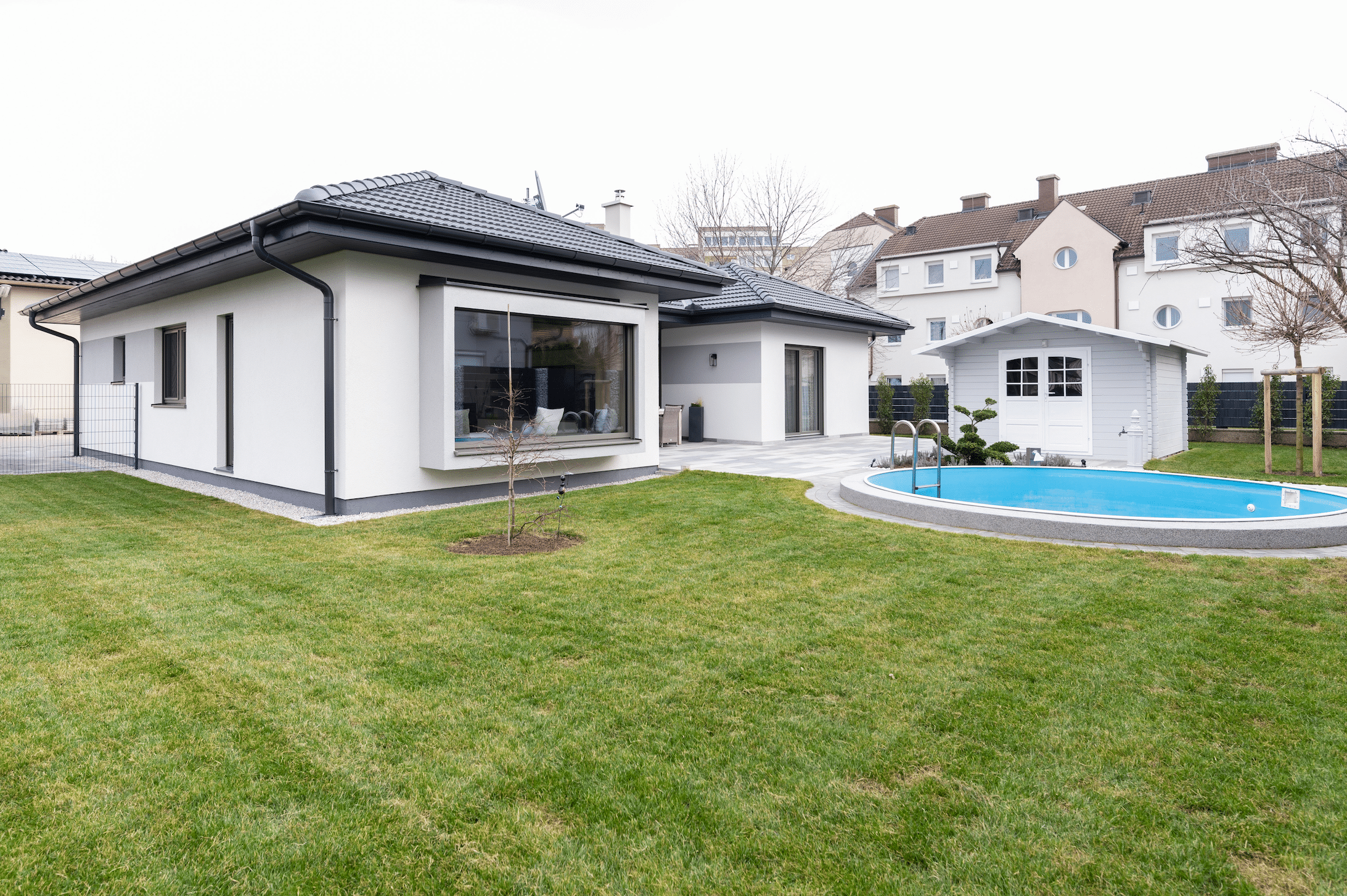 Hartl Haus zeigt einen weißen, modernen Bungalow mit schwarzem Walmdach, großen Fensterflächen, einen Pool und kleinem Gartenhäuschen.